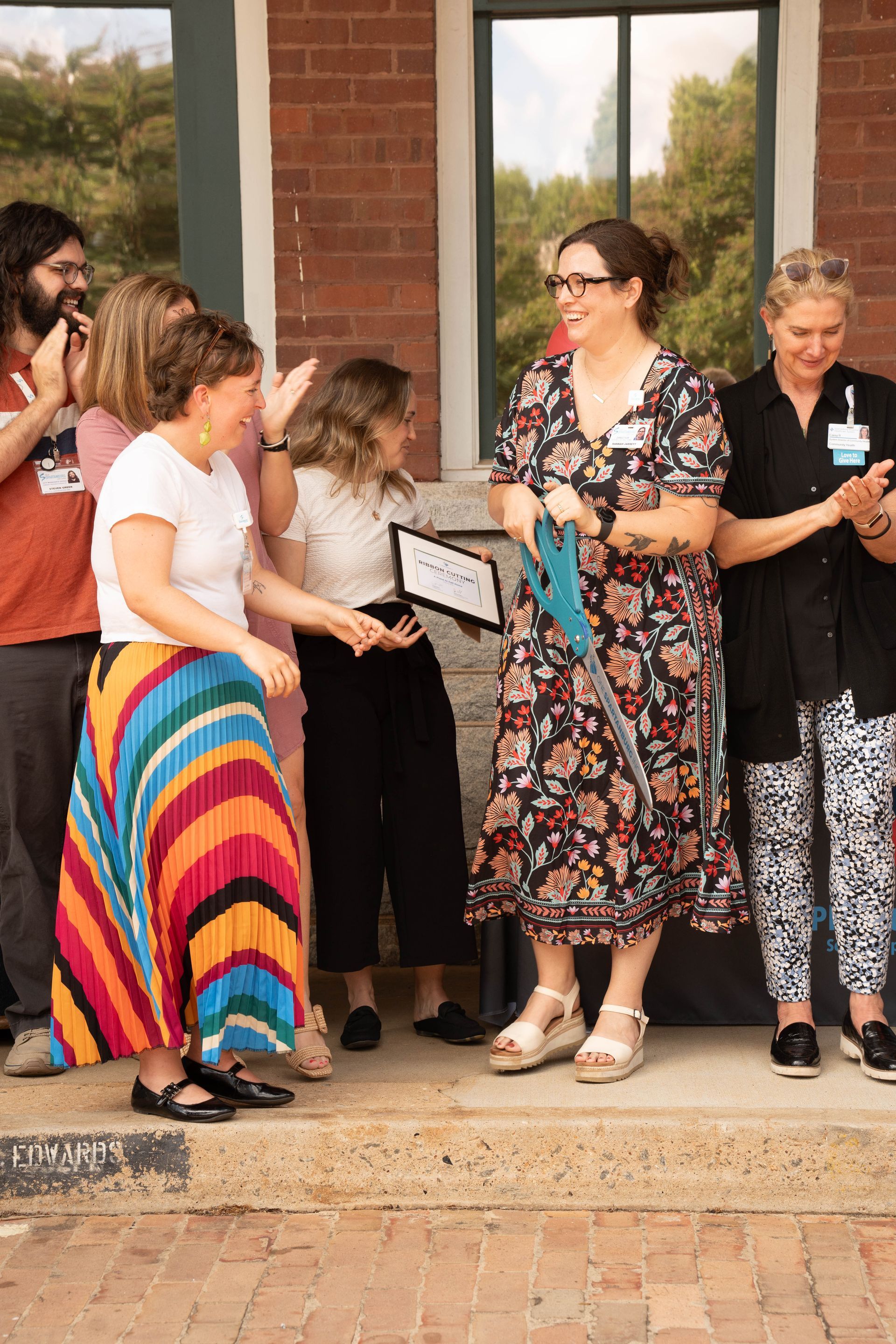 People celebrating a ribbon-cutting ceremony outside a brick building. A woman holds oversized scissors. Others clap and smile.