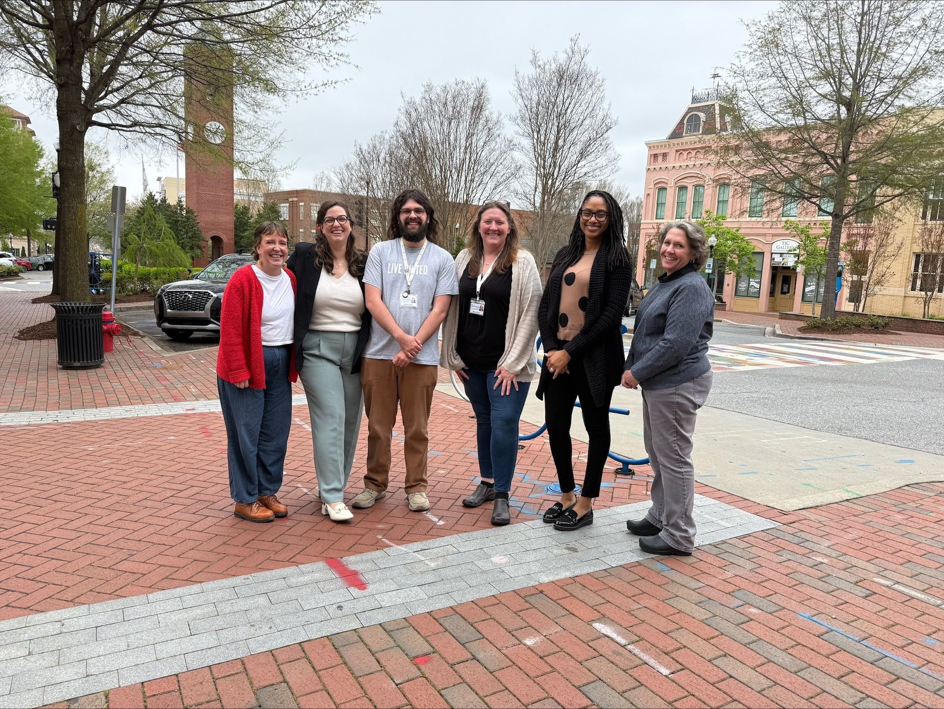 Group of six people standing on a brick plaza in front of a clock tower and buildings. Cloudy day.