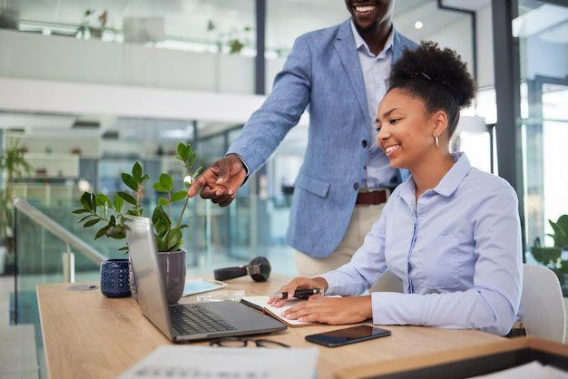 A man is standing next to a woman sitting at a desk with a laptop.