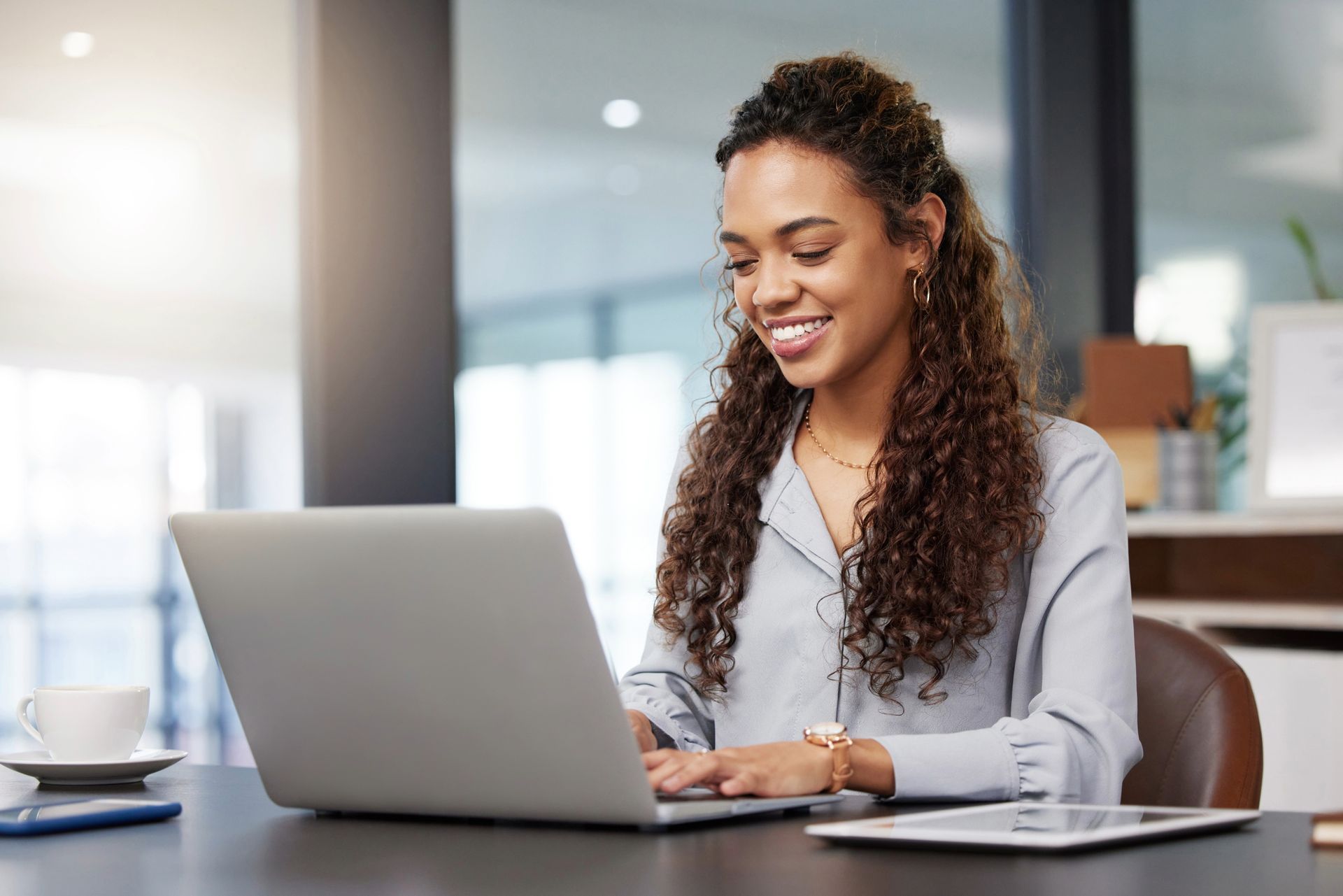 Woman smiling while working on a laptop at a desk in an office.