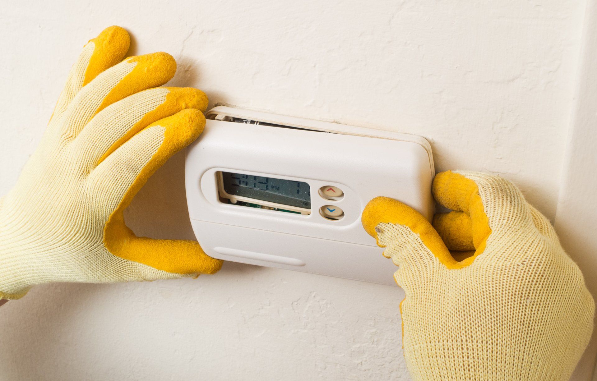 A person wearing yellow gloves is installing a thermostat on a wall.