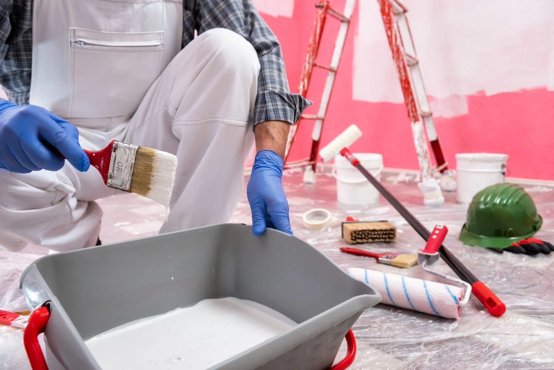 Person in white overalls painting a room with a paintbrush, tray, rollers, and drop cloths