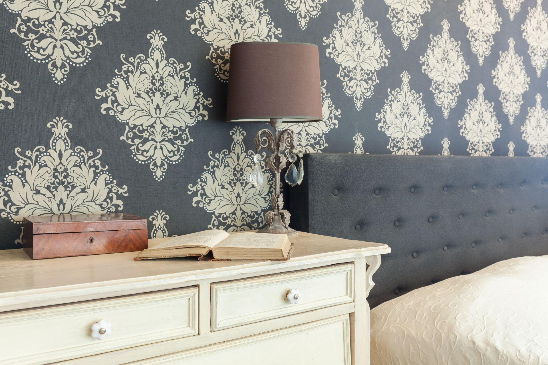 Bedroom corner with white dresser, patterned blue wallpaper, tufted gray headboard, and mauve lamp.