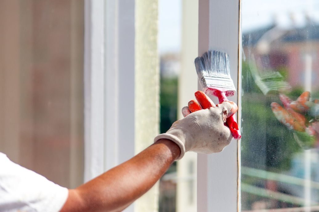 Gloved hand painting a white window frame with a red brush, next to a sunlit window