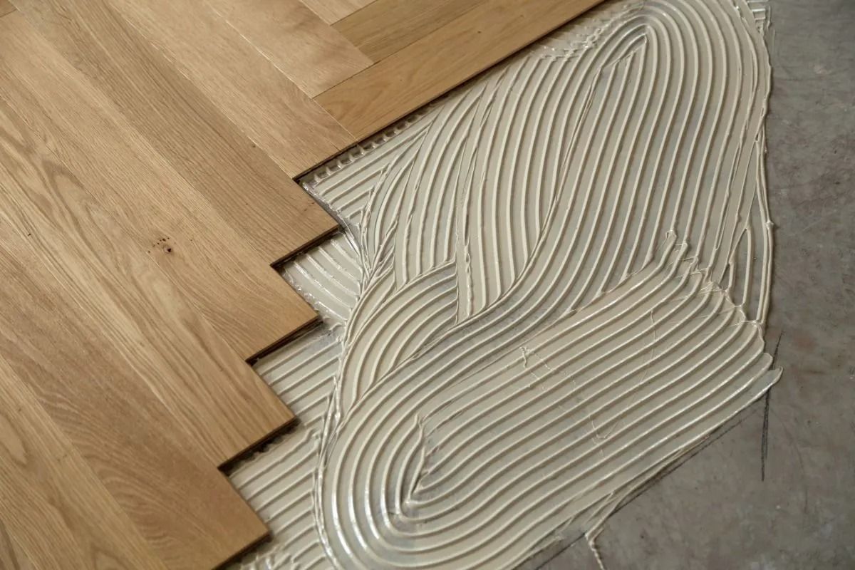 Wooden floor planks meeting gray adhesive spread with a notched trowel pattern