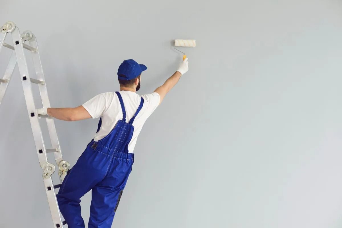 Painter on a ladder painting a light gray wall white with a roller