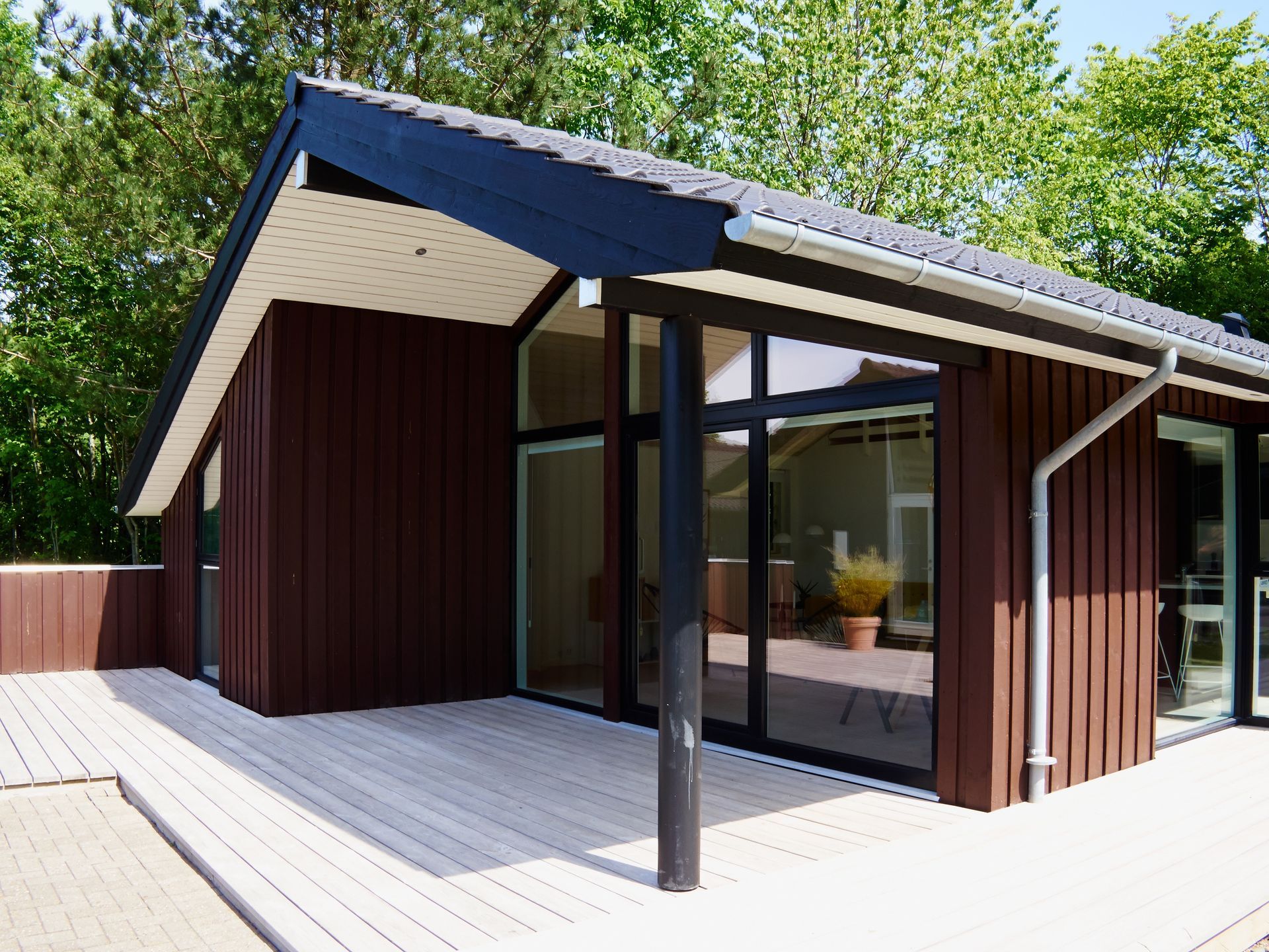 Cabane moderne en bois brun avec un toit à pignon, de grandes baies vitrées et une terrasse en bois clair, située en pleine forêt.