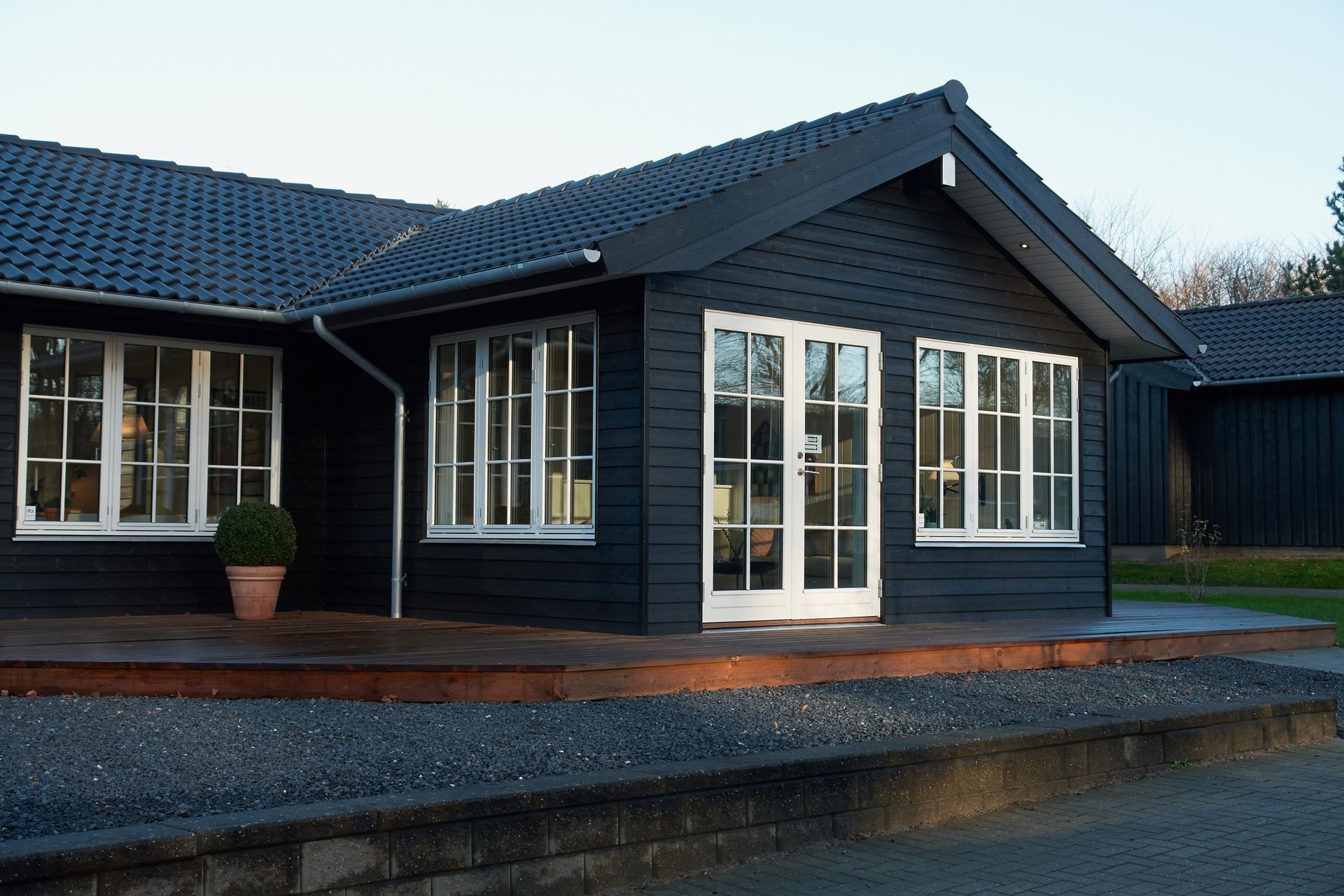 Une maison de plain-pied en bois noir avec des fenêtres à cadre blanc et des portes-fenêtres doubles, comprenant une terrasse en bois et une cour en gravier.