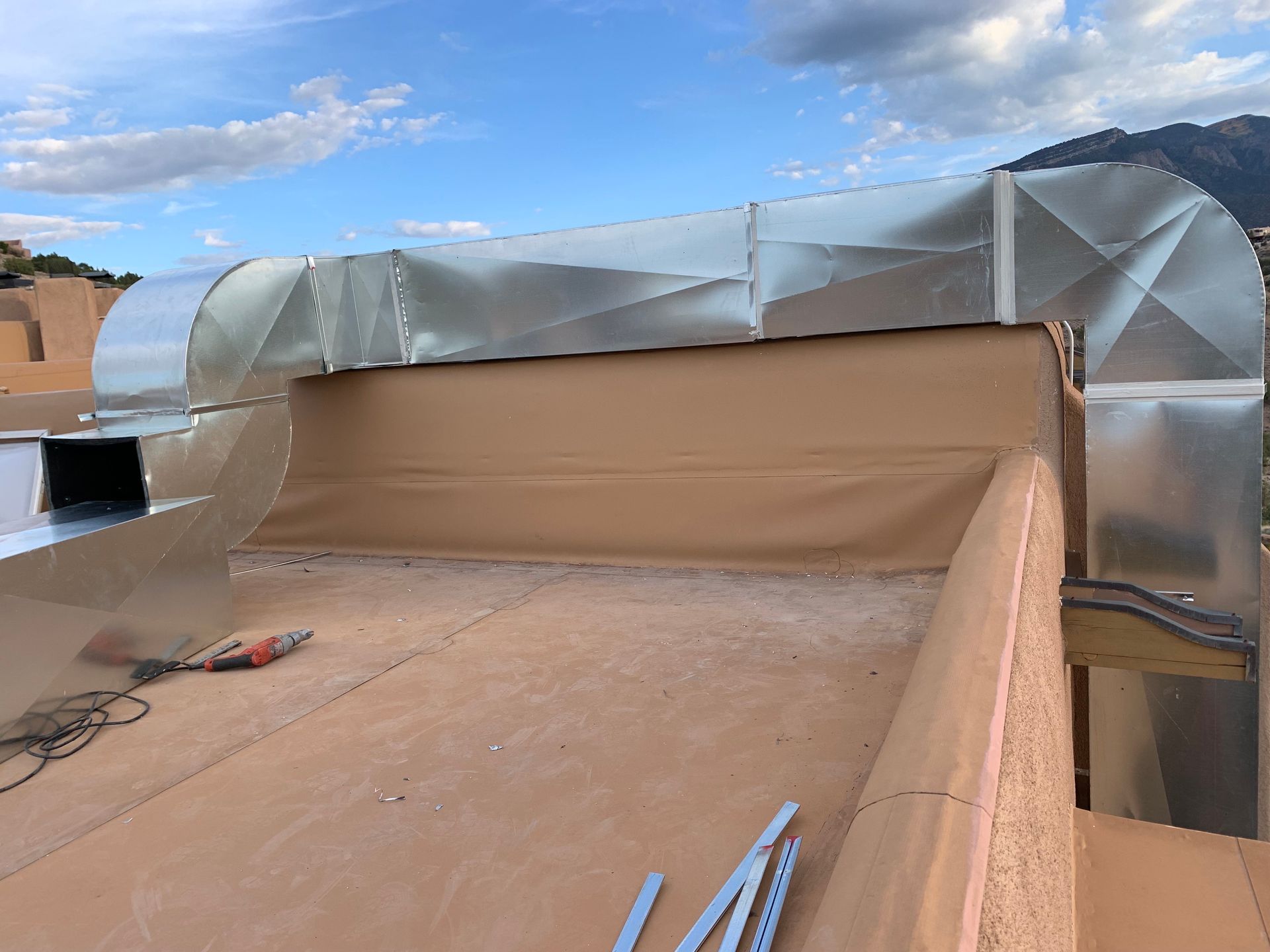 Rooftop with air ducts, tan stucco, and a mountain backdrop. Tools and metal brackets are visible.
