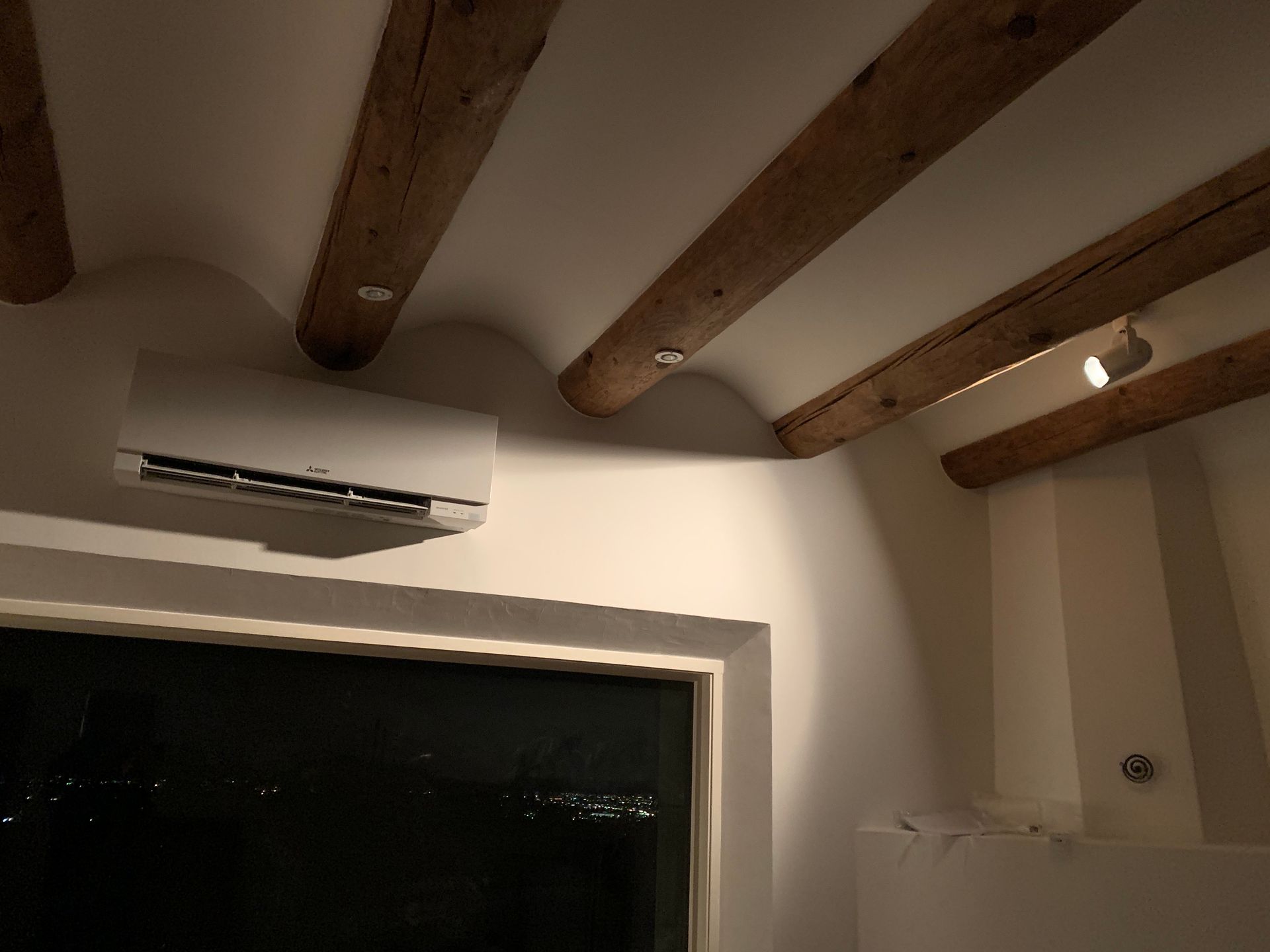 Air conditioner on white wall with wooden beams and a window overlooking a dark scene.