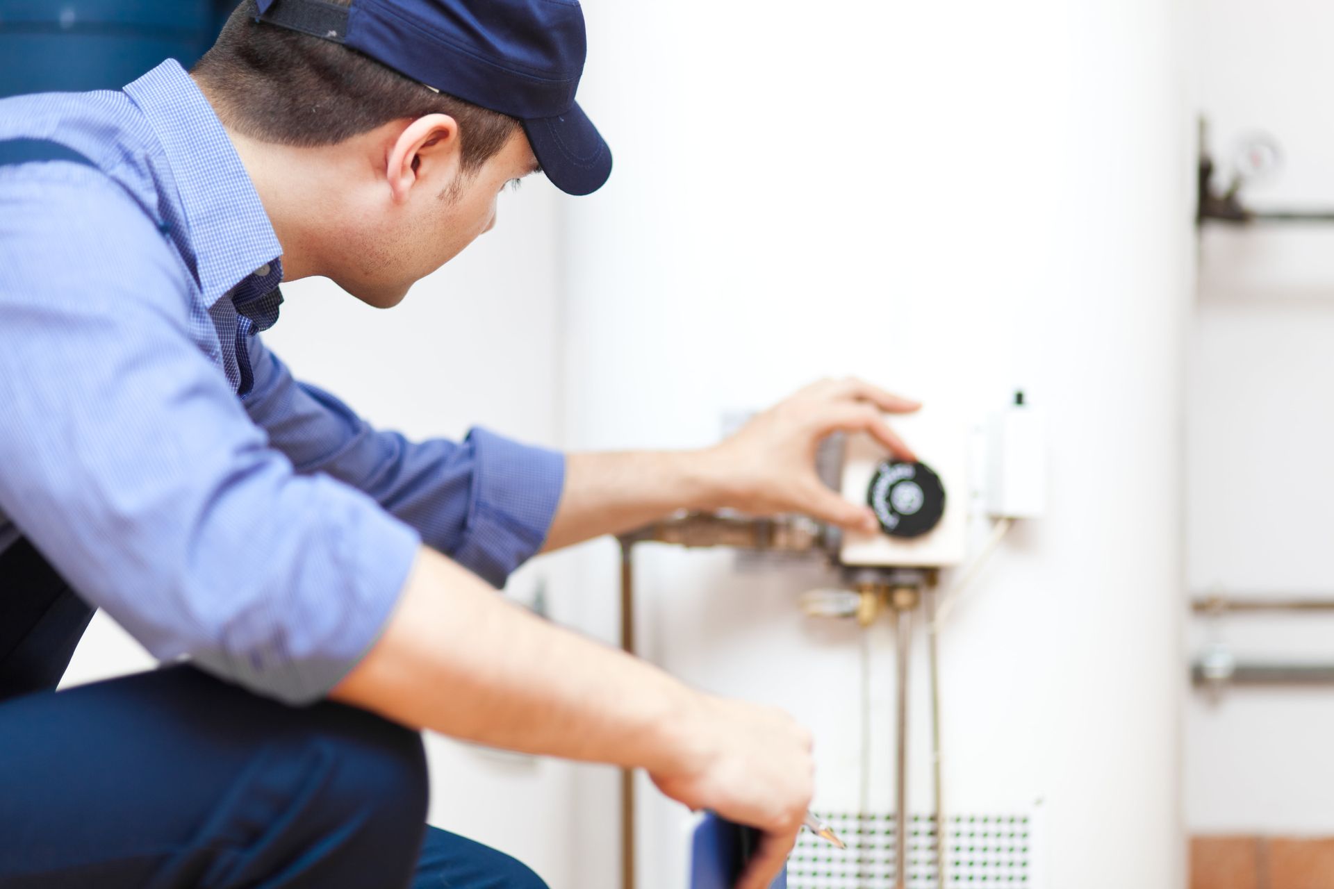 Plumber in blue shirt and cap adjusting a water heater's temperature control in a utility room.