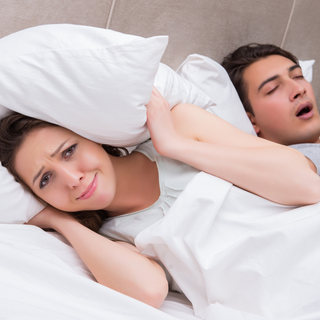 A man and woman are laying in bed. The woman covers her head with a pillow as the man snores.