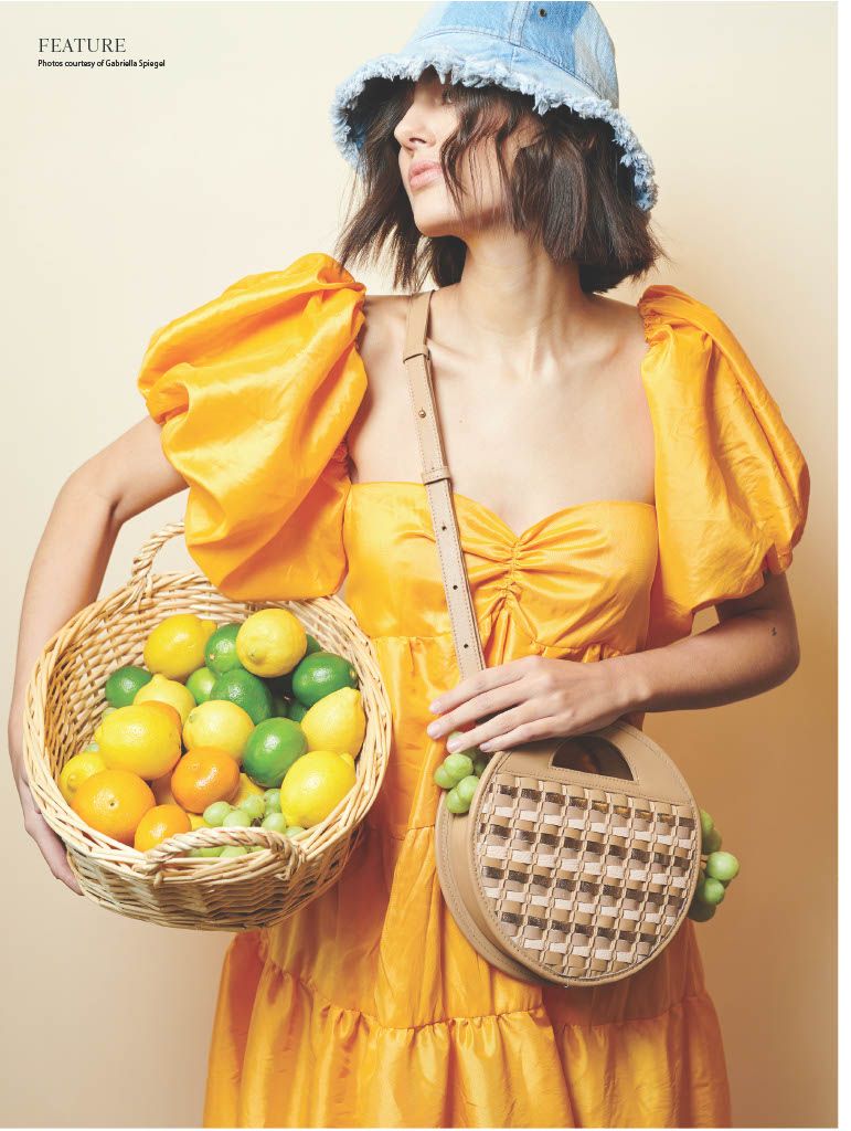 A woman in an orange dress, wearing a blue denim bucket hat, holding a basket of lemons and limes on her left hip