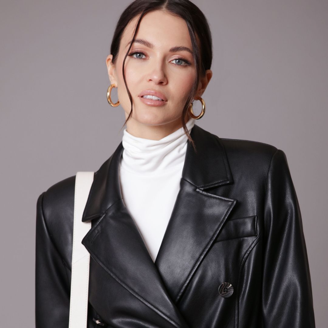 A woman wearing a ponytail, gold hoop earrings, a black leather trenchcoat, a white turtleneck underneath with a white bag strap on her left shoulder