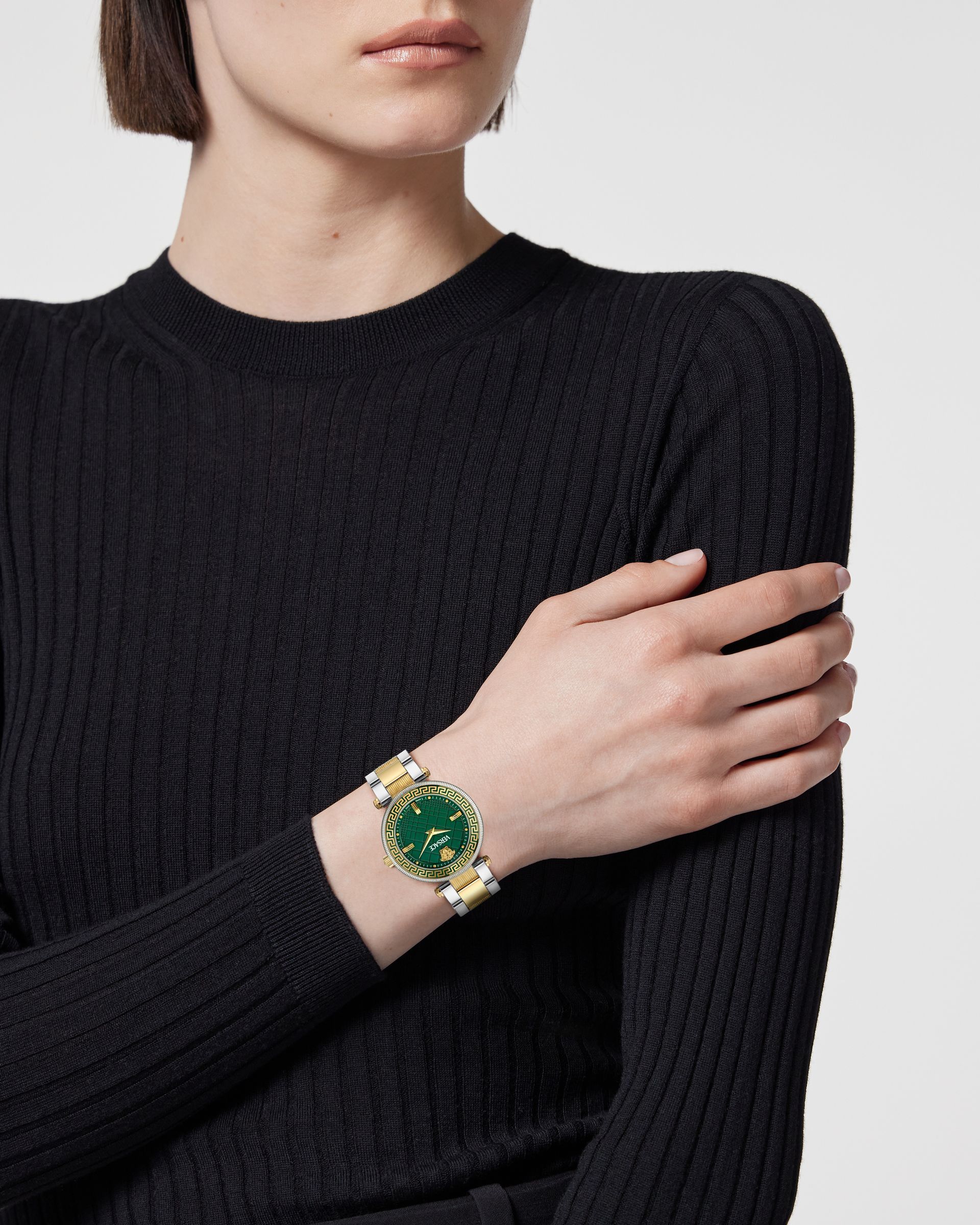 A woman with a short brunette bob in a black long-sleeved shirt with her hand on her right arm, showing a green-faced watch on her wrist