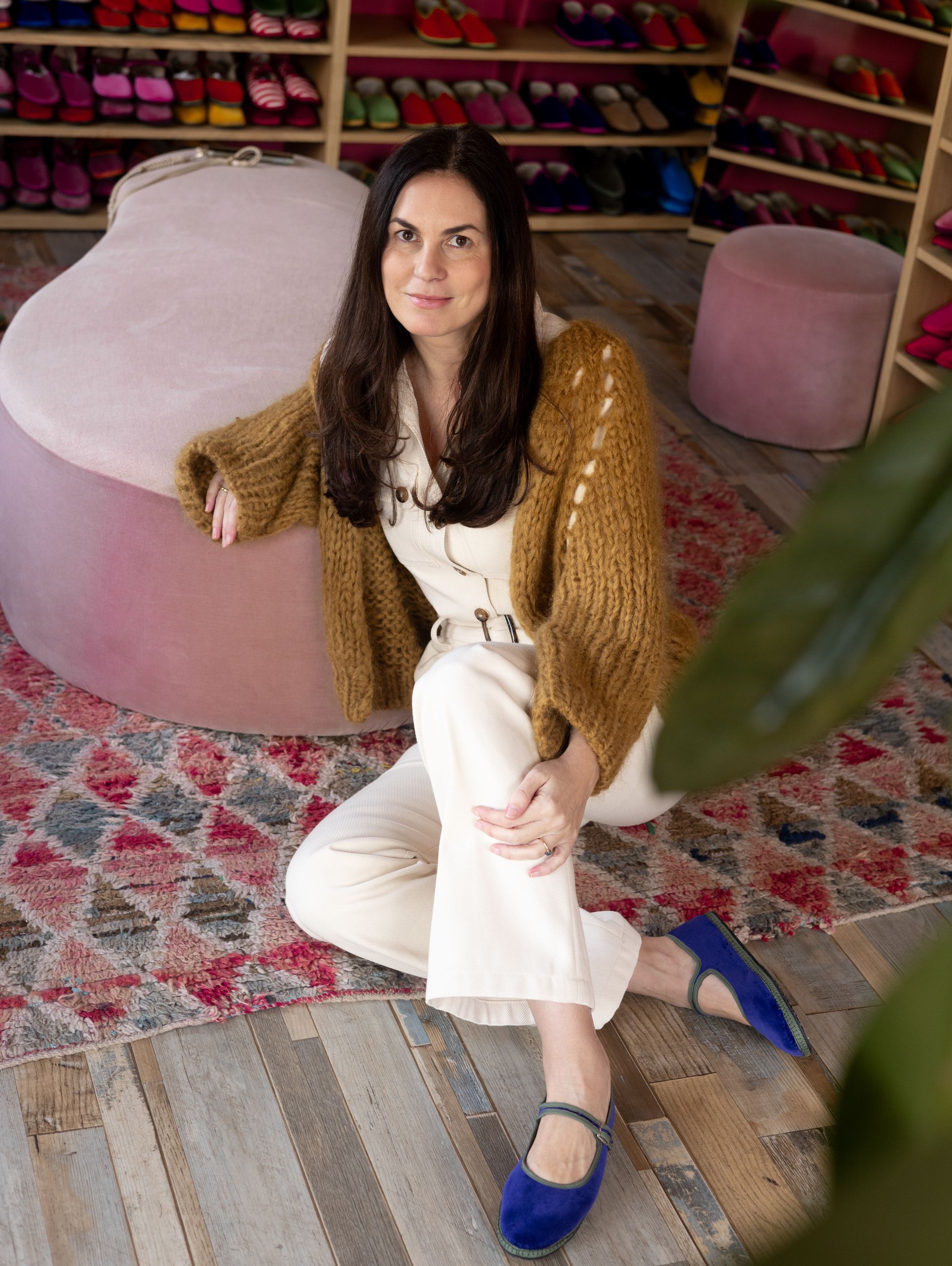 A brown-haired woman sitting on the floor, wearing a knitted yellow-brown cardigan, white pants, and royal blue velvet friulane flat shoes