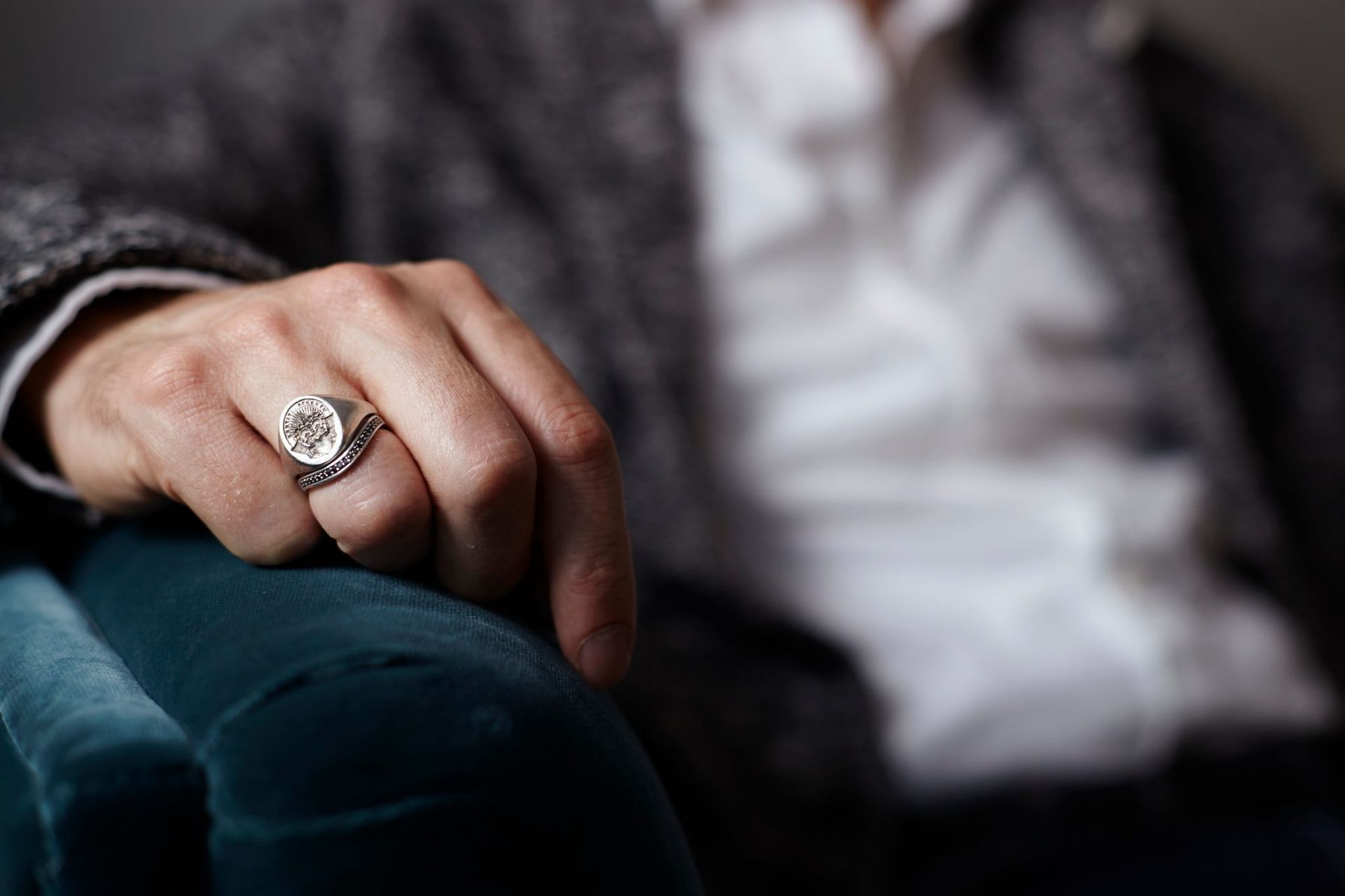 A hand's hand on a dark blue couch armrest wearing a silver signet ring on his ring finger