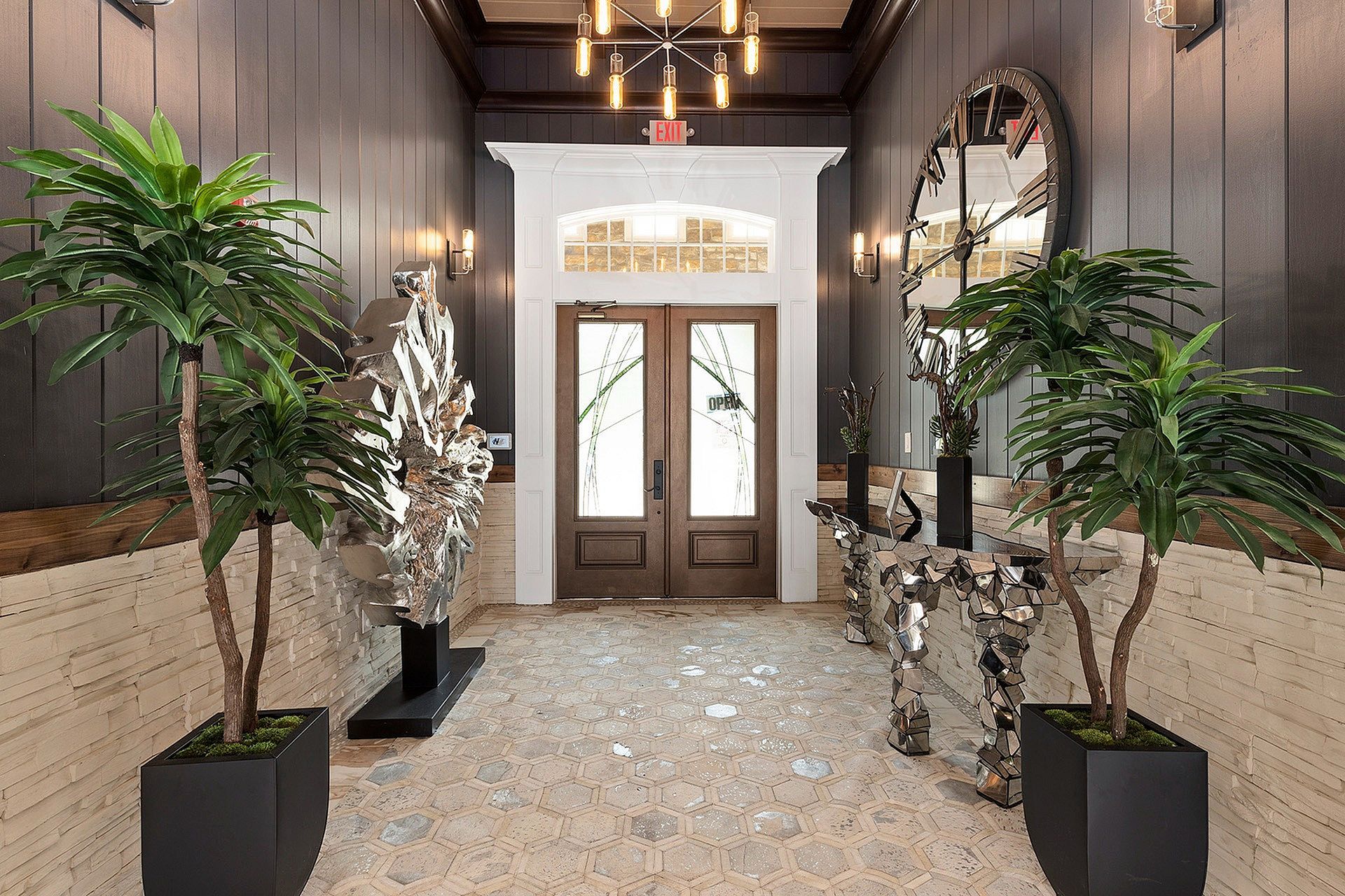 Lobby entrance with double wooden doors, pendant chandelier, and large potted plants.