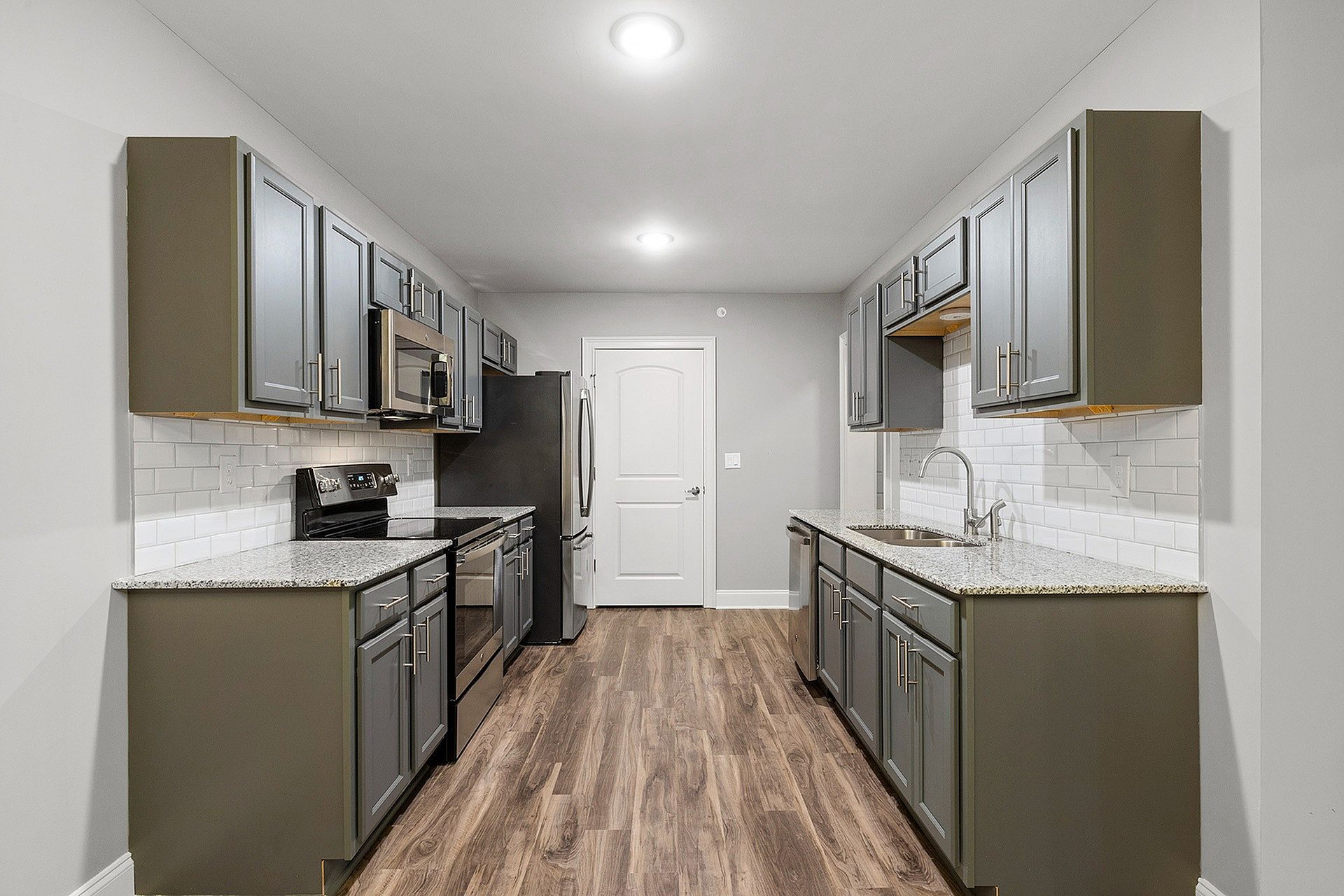 Galley kitchen with gray cabinets, granite countertops, white subway tile, and stainless steel appliances.