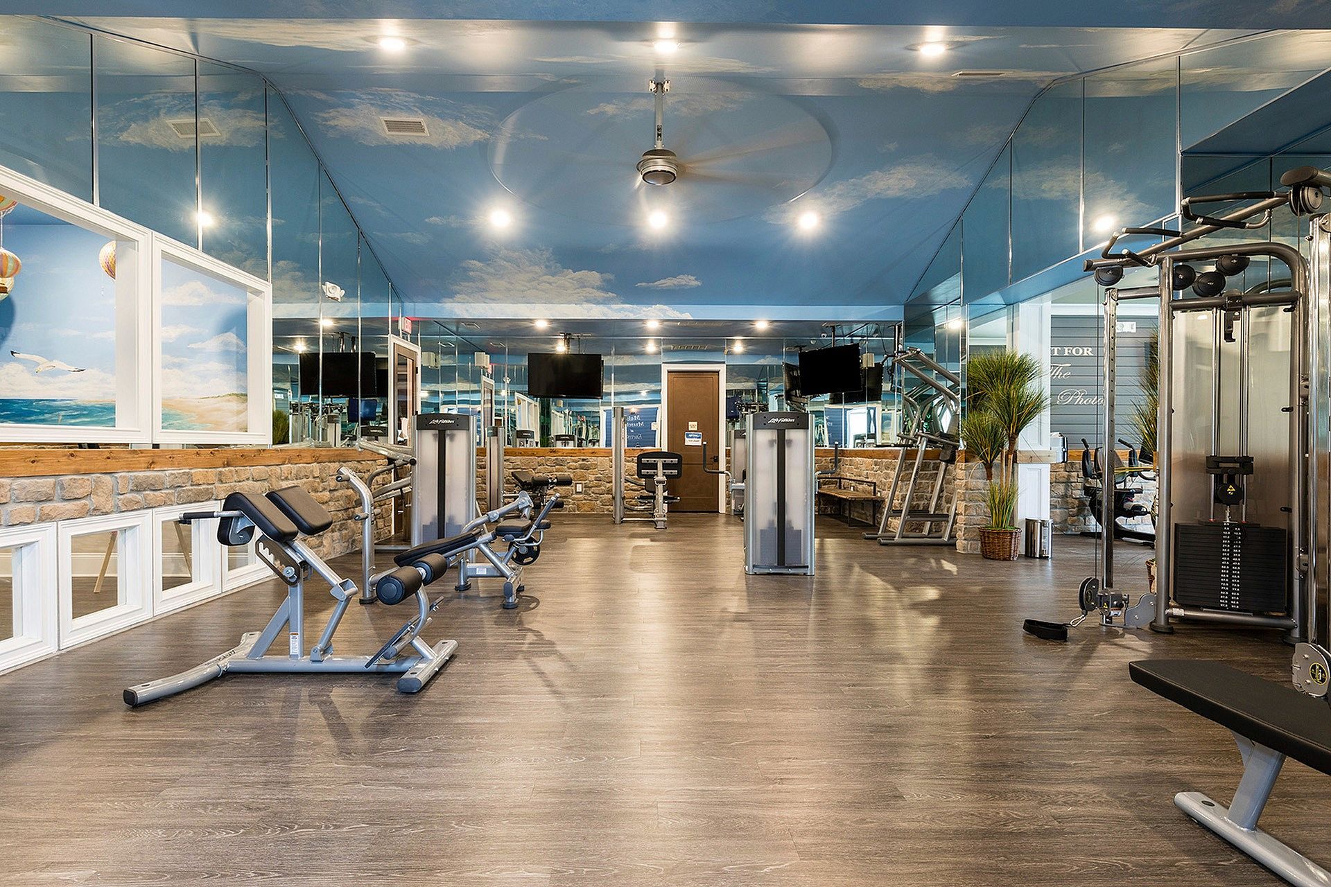 Bright gym with multiple workout machines, mirrors, and a blue sky ceiling.