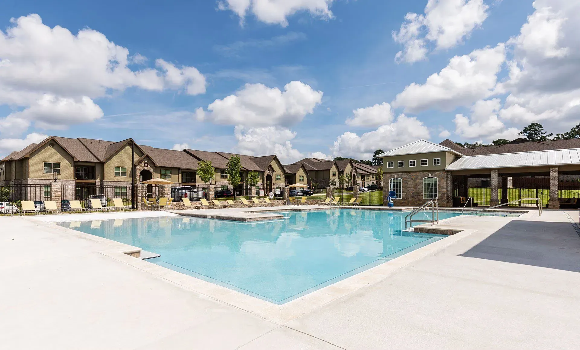 Swimming pool and lounge chairs at apartment complex, with blue sky and clouds.