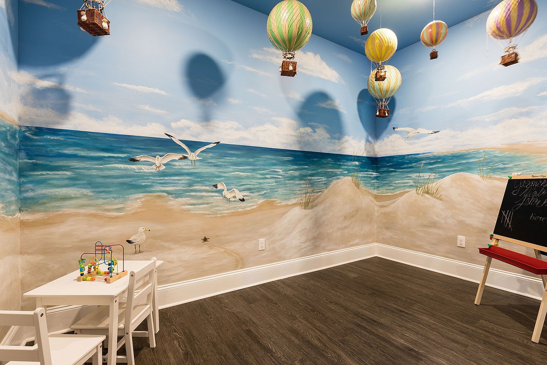 Children's playroom with a beach mural, hanging hot-air balloon decorations, and kid-sized table and chairs.