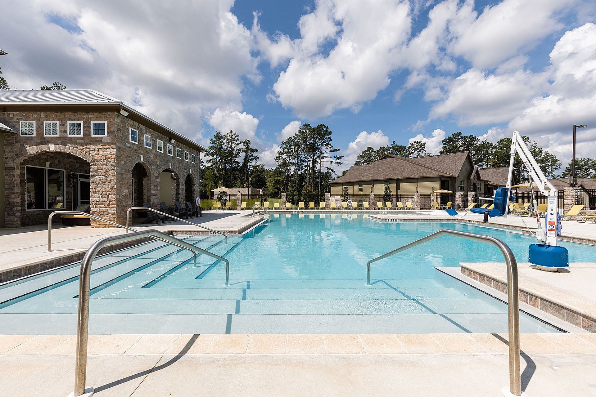 Outdoor community pool with steps, metal handrails, and a pool lift beside a stone clubhouse.