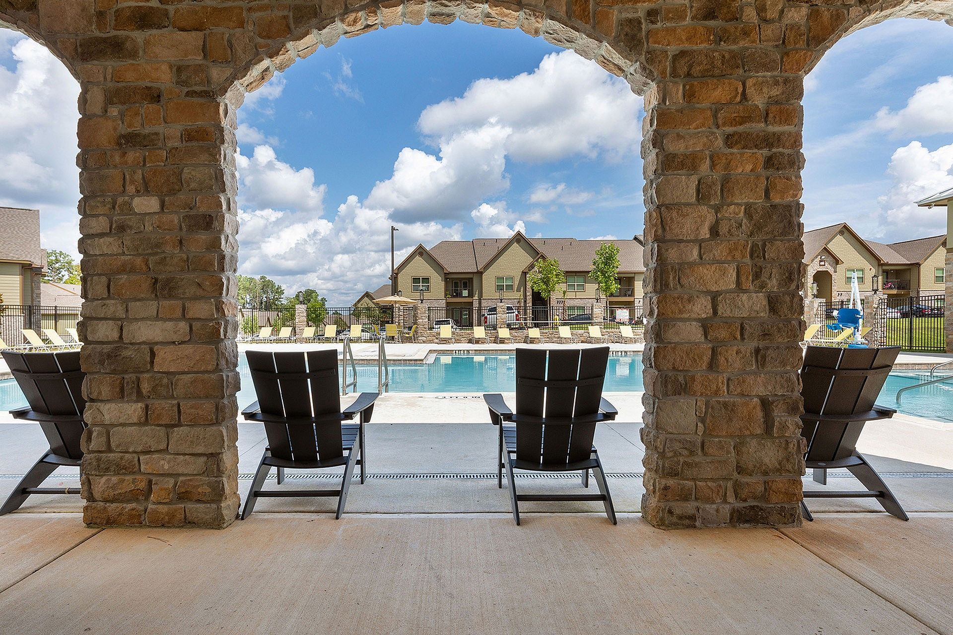 Stone archway opening to a community pool with lounge chairs and apartment buildings beyond.
