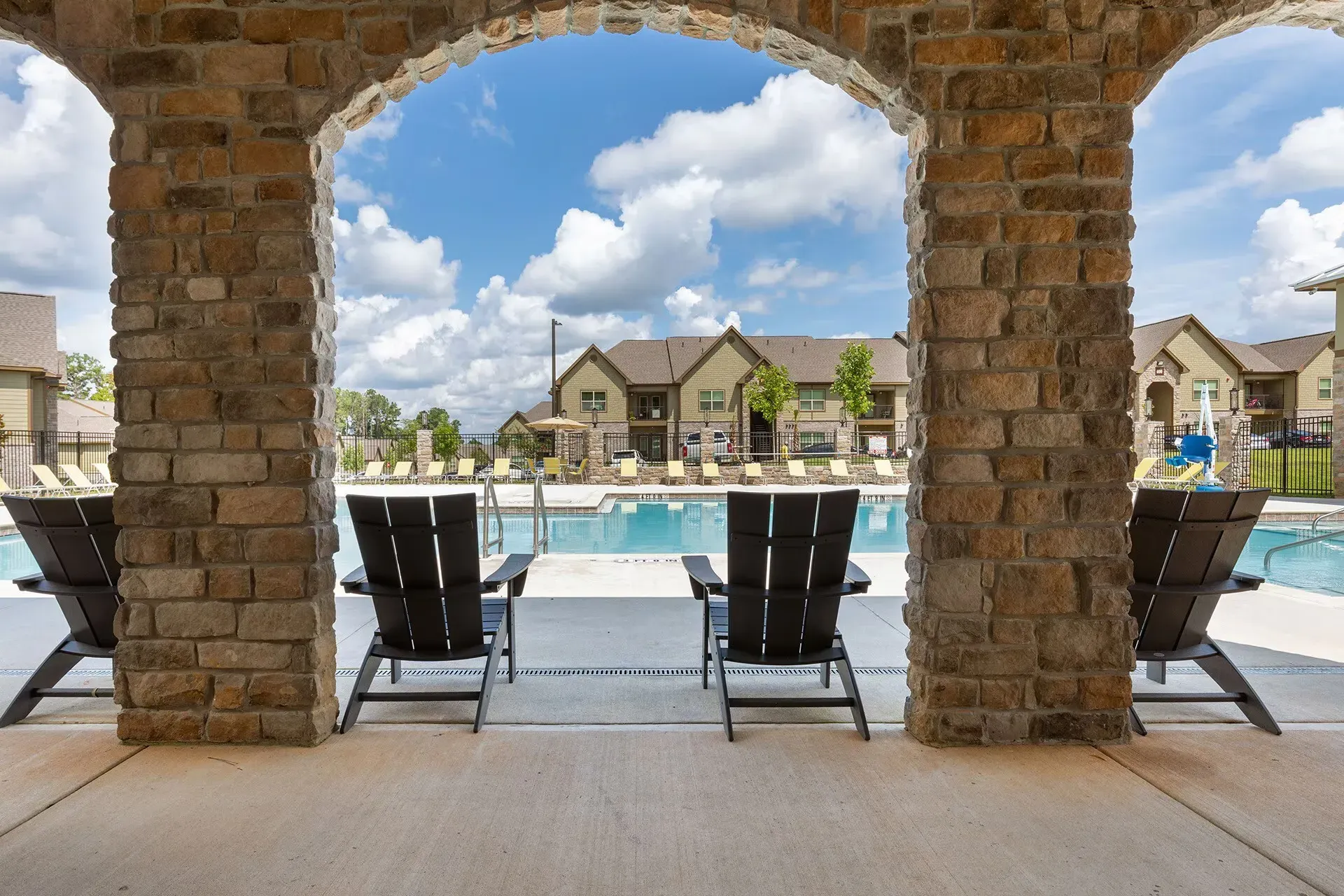 Stone archway frames a pool with lounge chairs, overlooking apartment buildings under a blue sky.