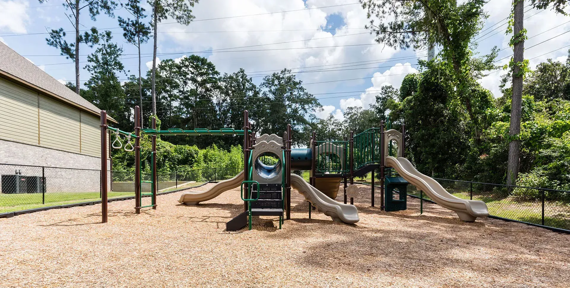 Playground with slides, climbing structures, and swing set, set on wood chips, with trees in background.