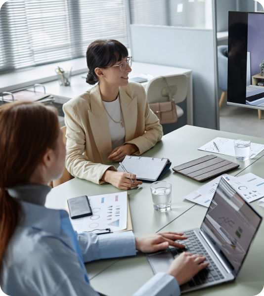 Three coworkers in a bright office meeting, reviewing documents and a laptop at a glass table.