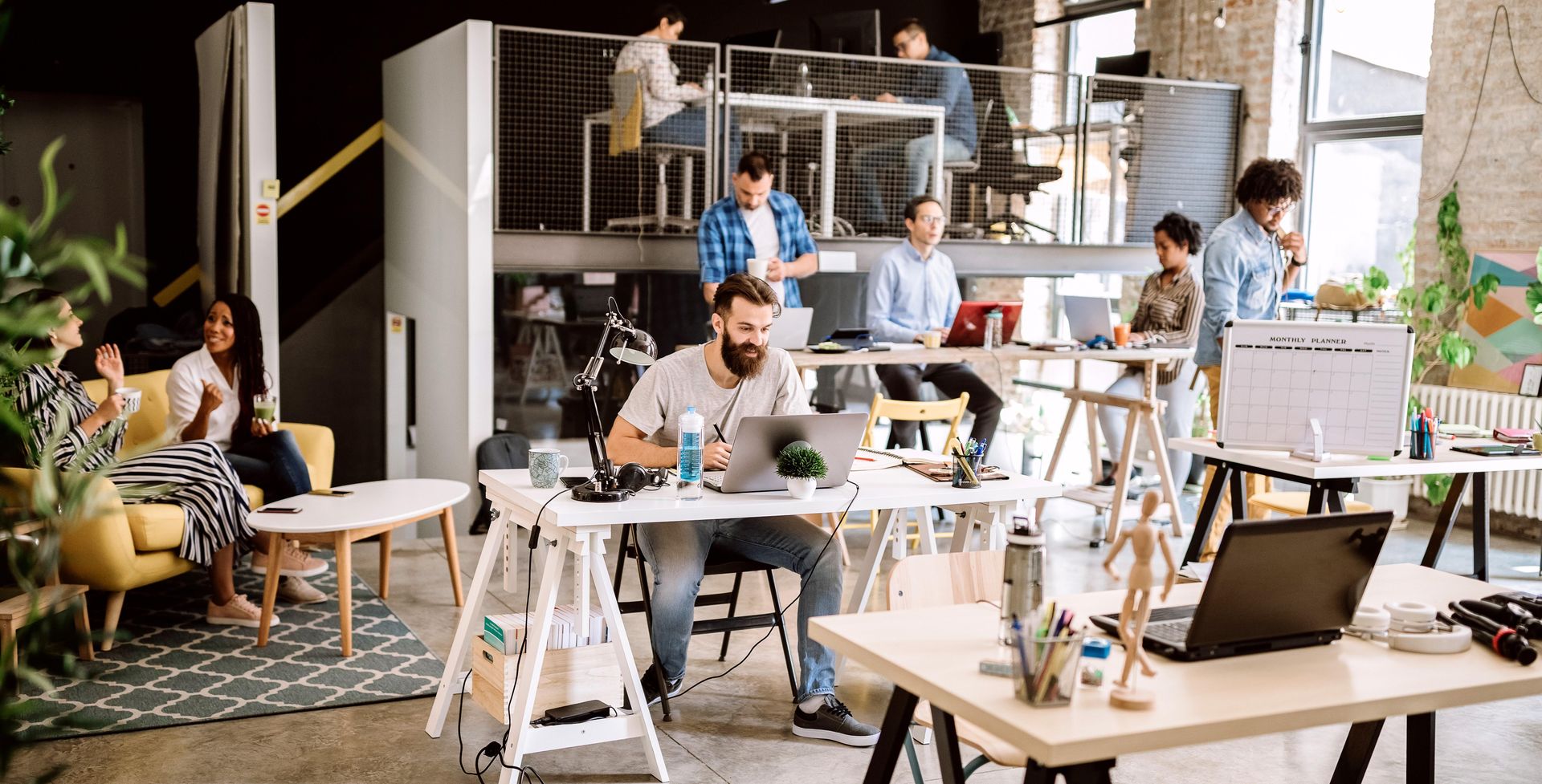 Open coworking space with people working at tables and laptops, lit by large windows
