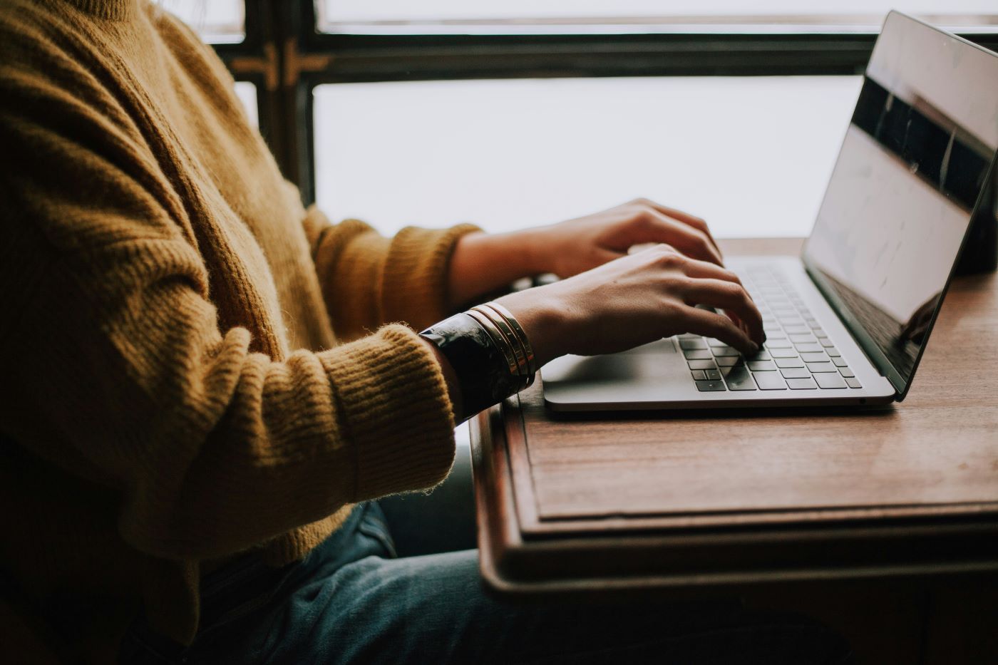 Person typing on a laptop at a wooden table near a window, wearing a yellow sweater and jeans.