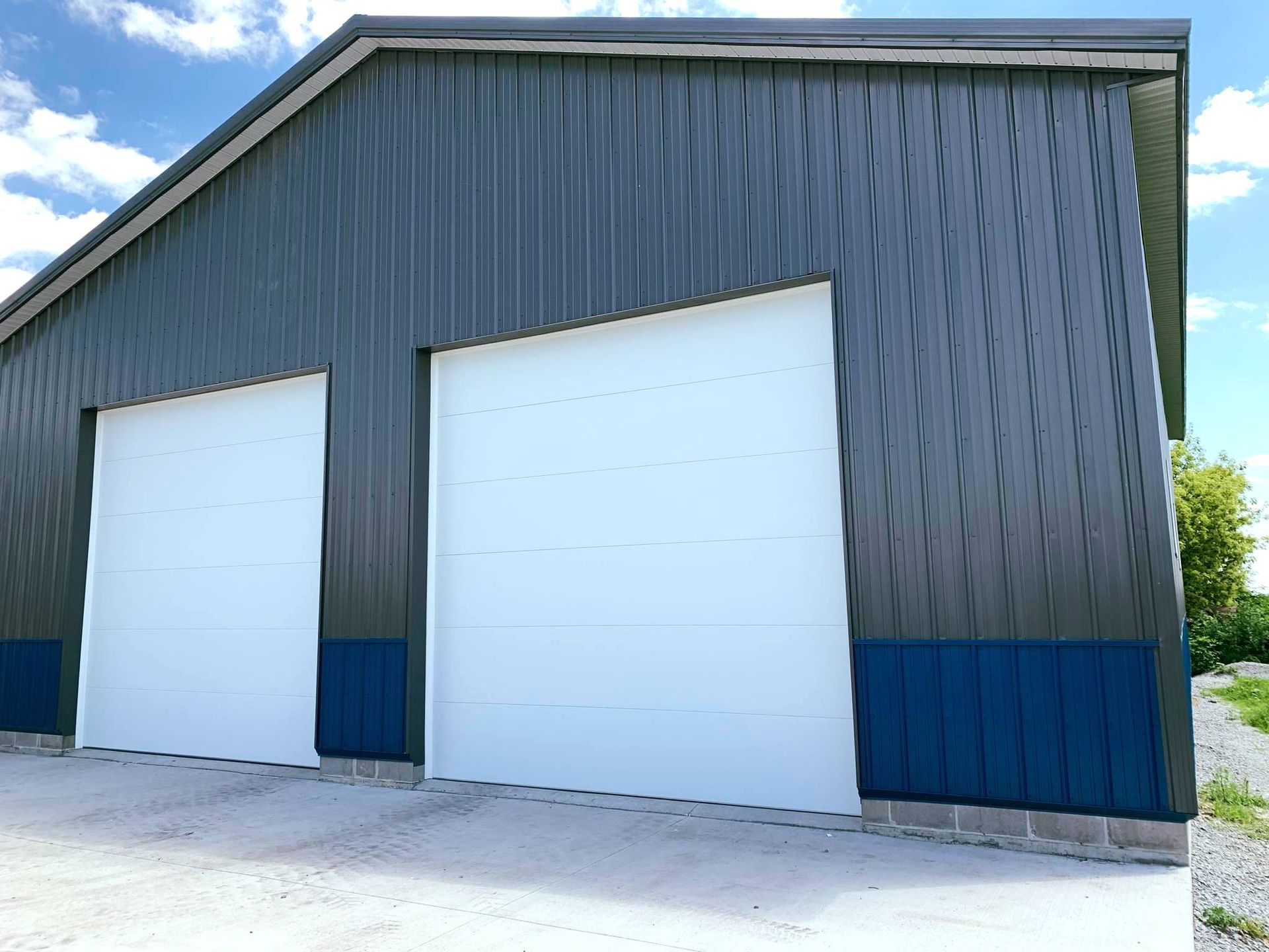 Two-bay steel building with white garage doors, dark blue and gray exterior against a partly cloudy sky.