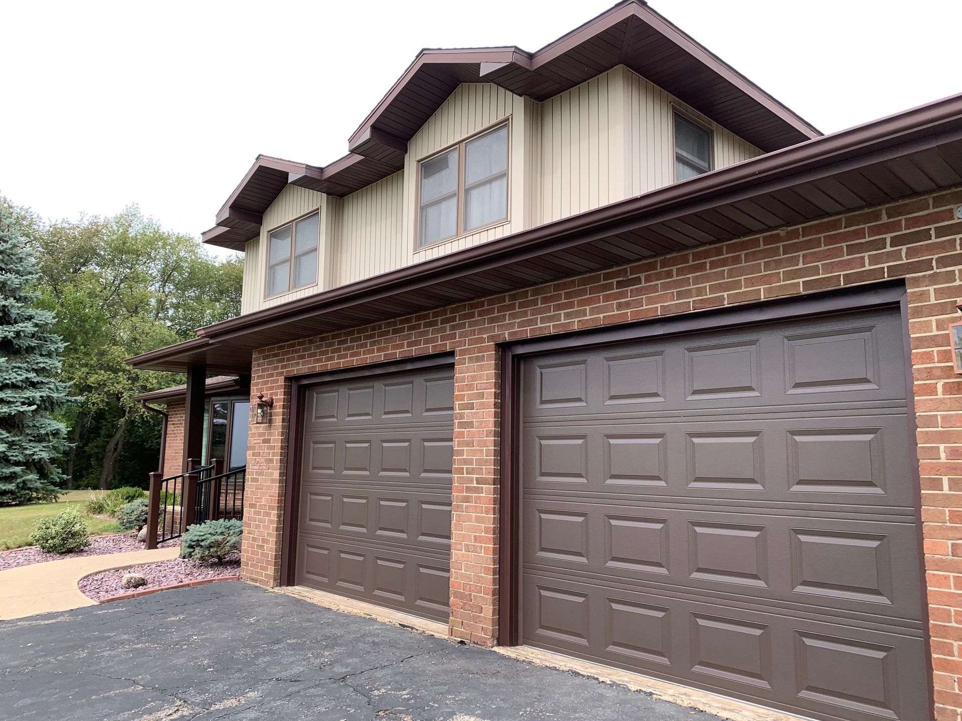 Two-story house with brown garage doors, brick, and tan siding, cloudy sky.