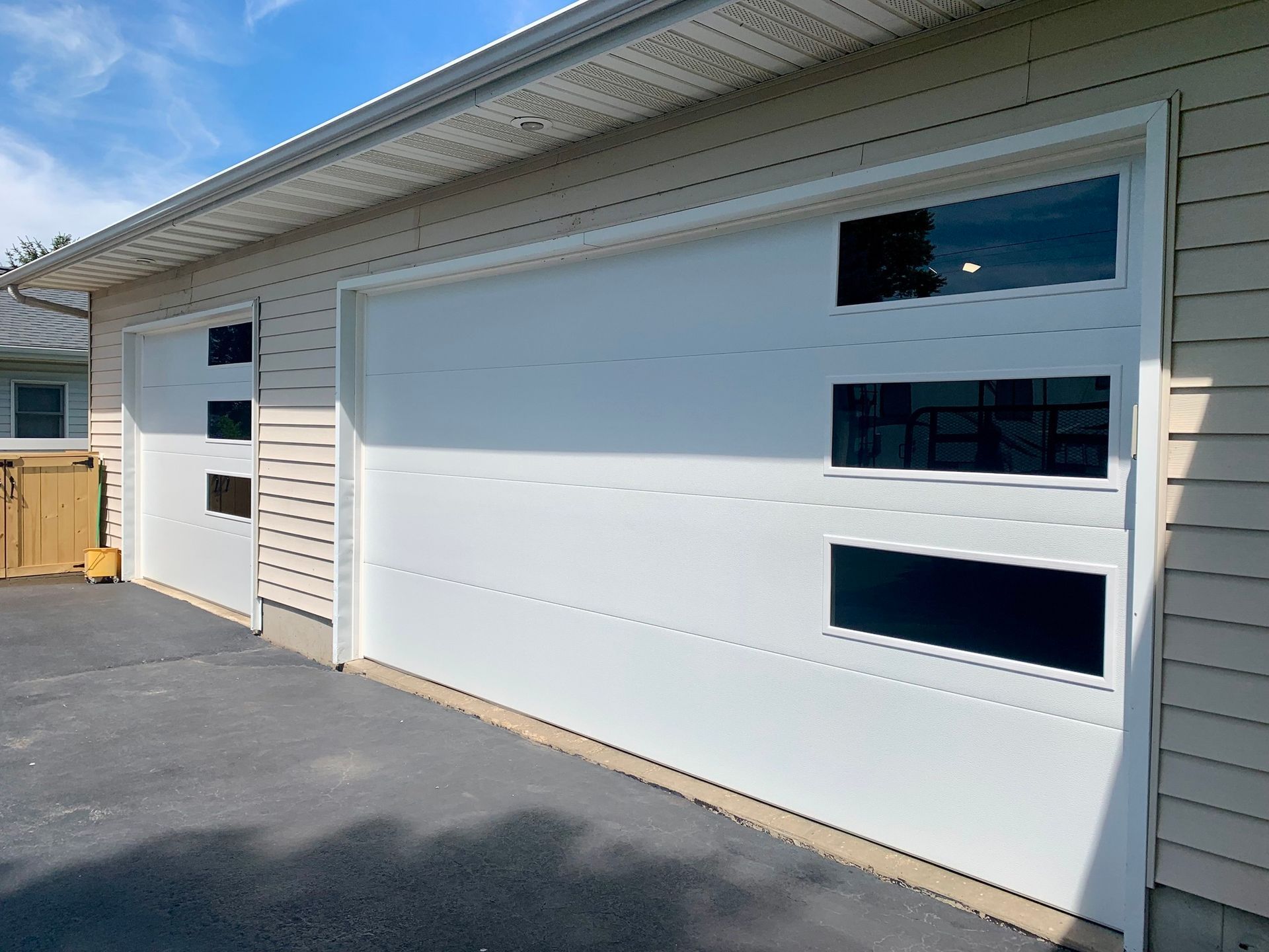 White garage doors with black windows, on a building with tan siding, a black driveway.