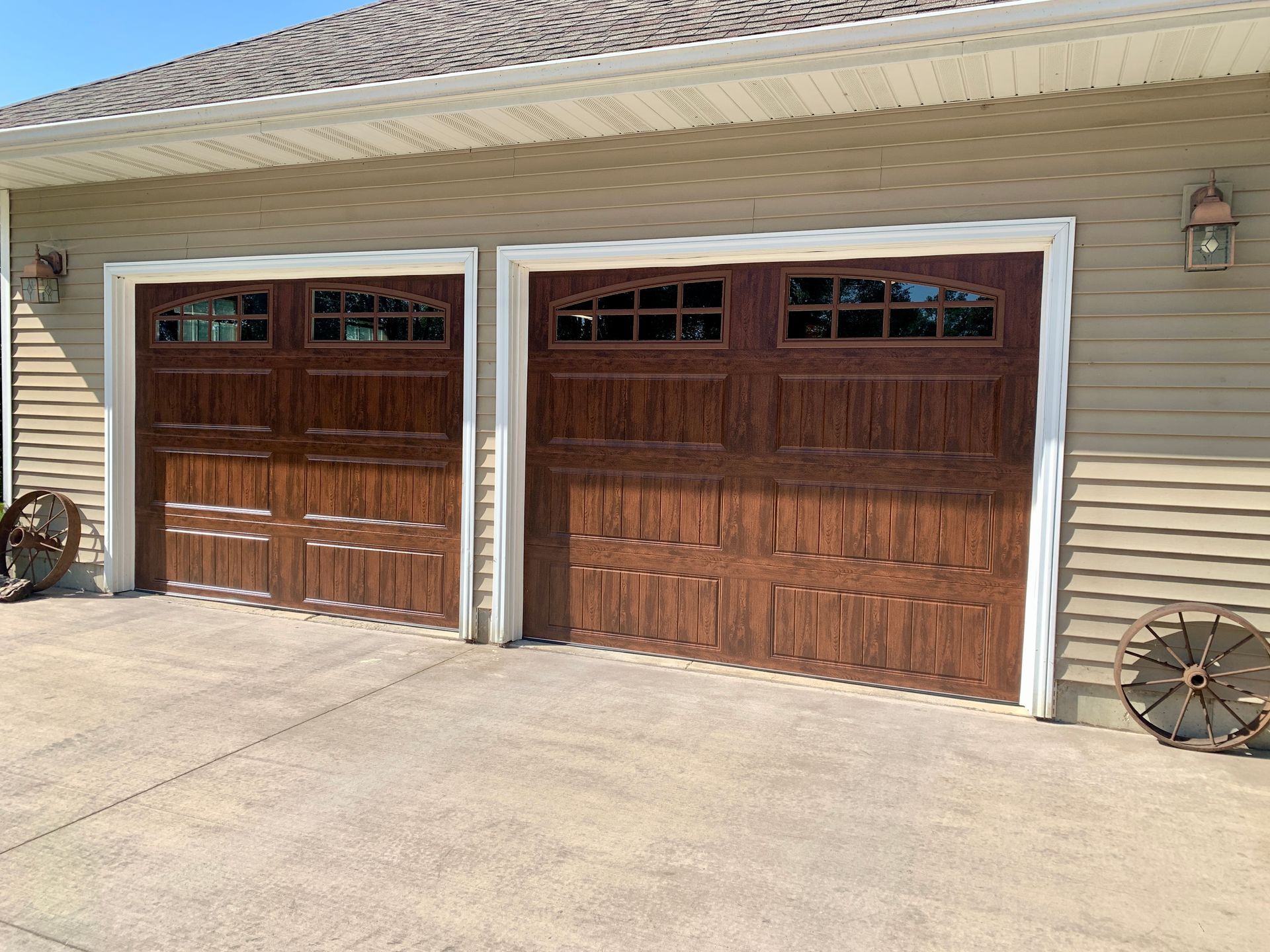 Two brown garage doors with glass windows in a tan-colored building with a concrete driveway.
