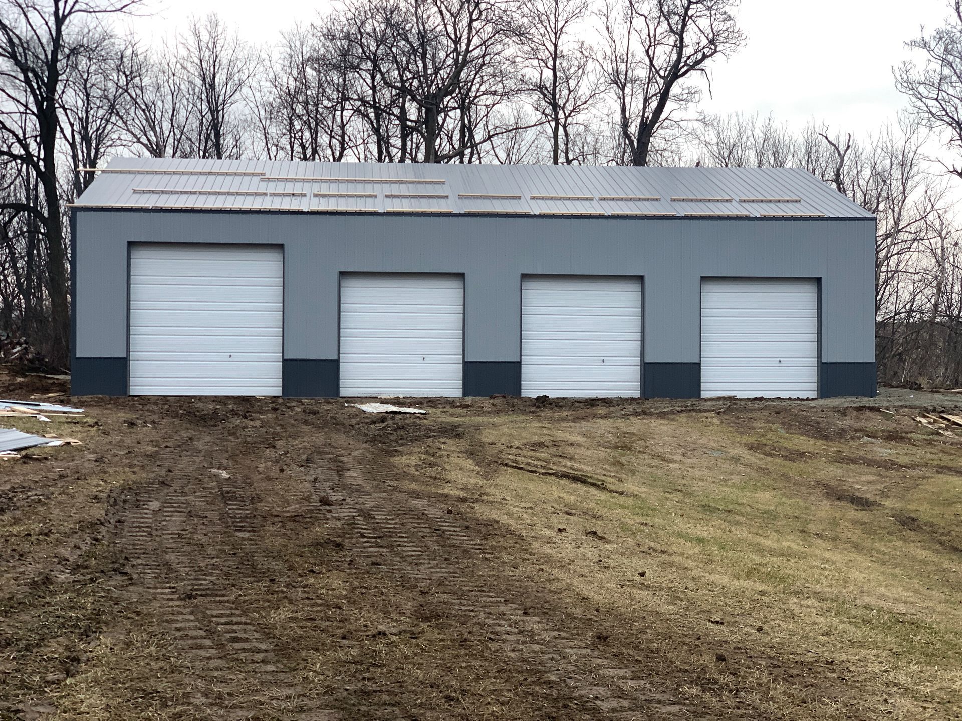 Gray garage with four white garage doors, set on a muddy hill with bare trees in the background.