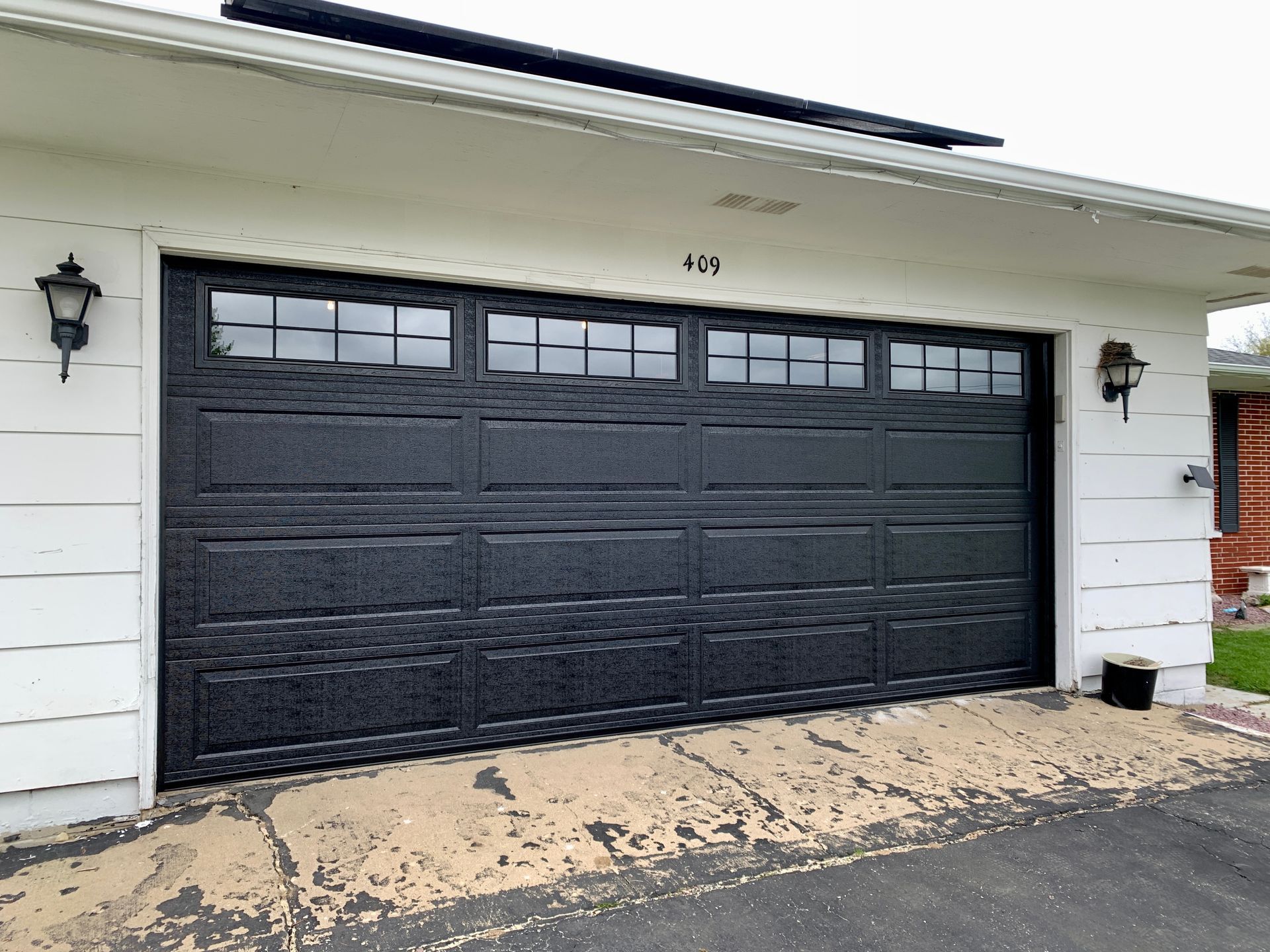 Black garage door with windows, set in a white house, with two sconces.