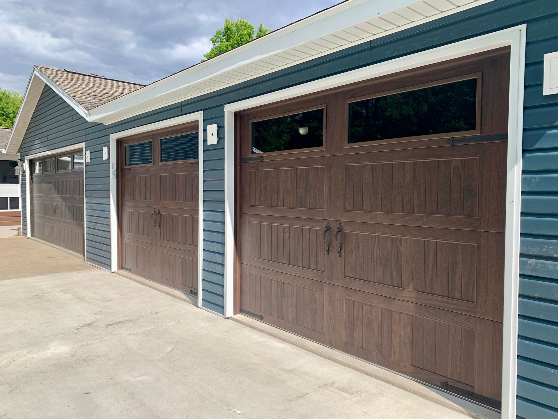 Three brown garage doors with windows on a blue house with white trim. Concrete driveway.