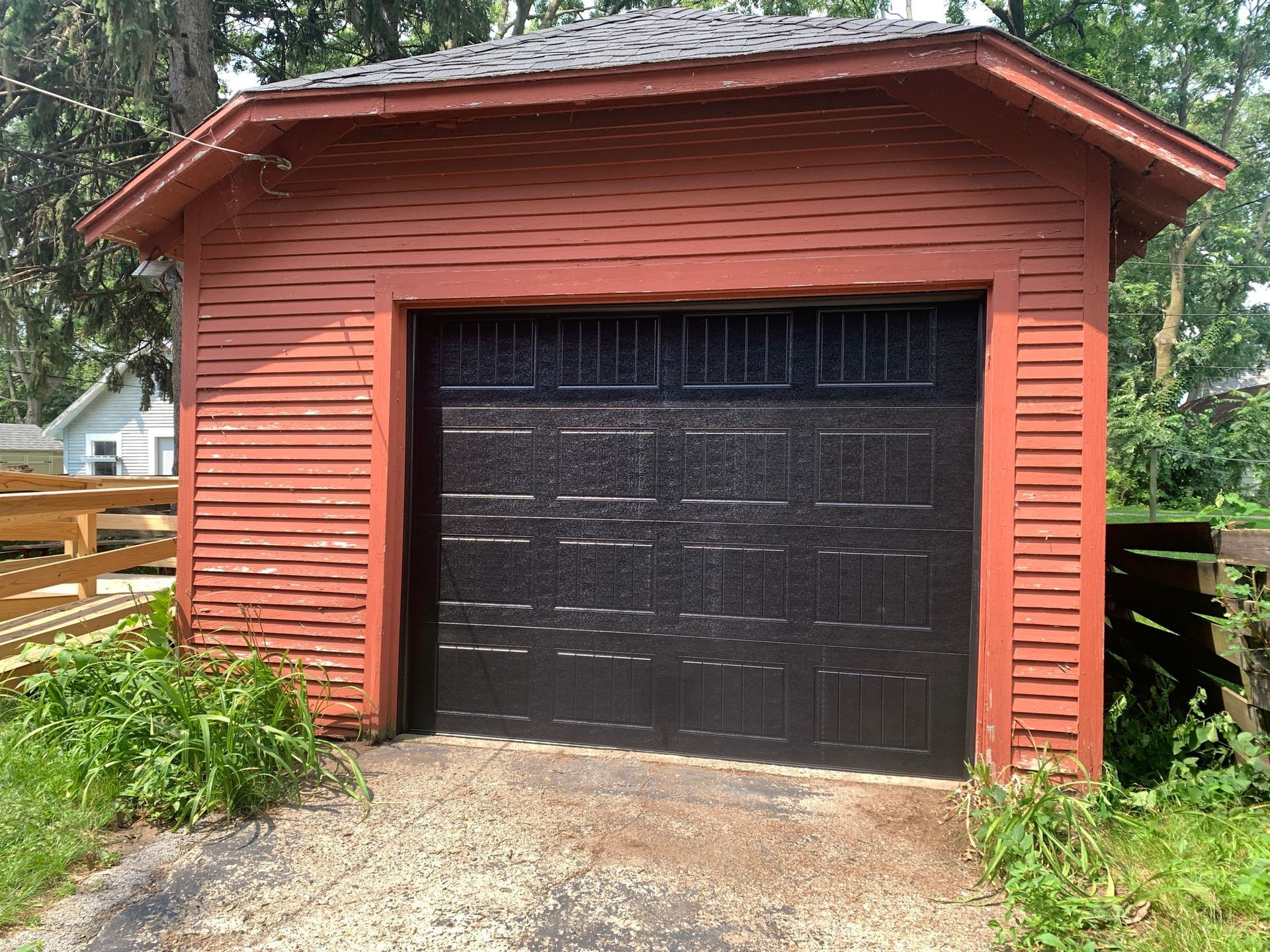 Red wooden garage with black door. Gravel driveway, green plants.