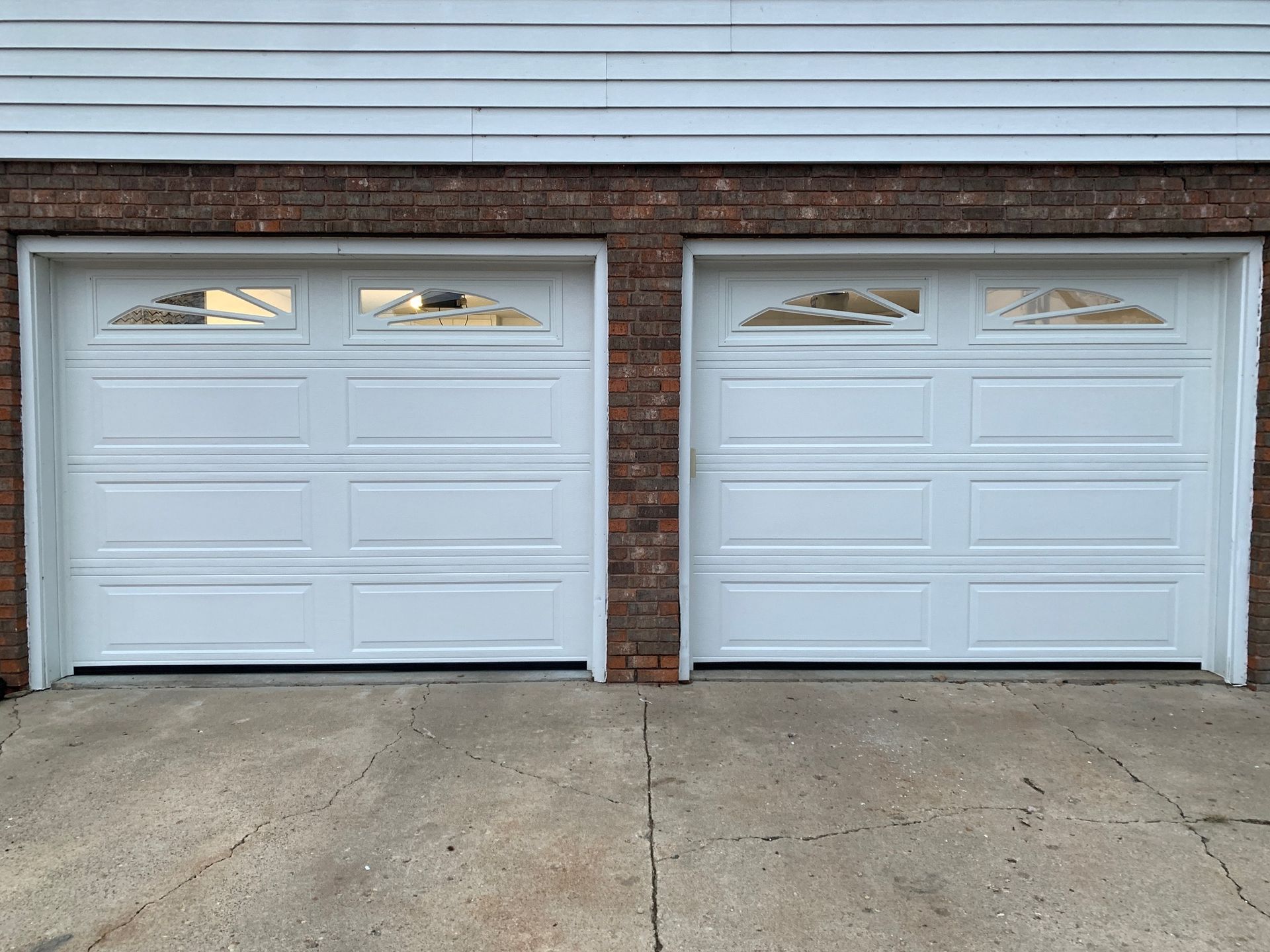 Two white garage doors with glass panels, set in a brick and siding building, on a concrete driveway.