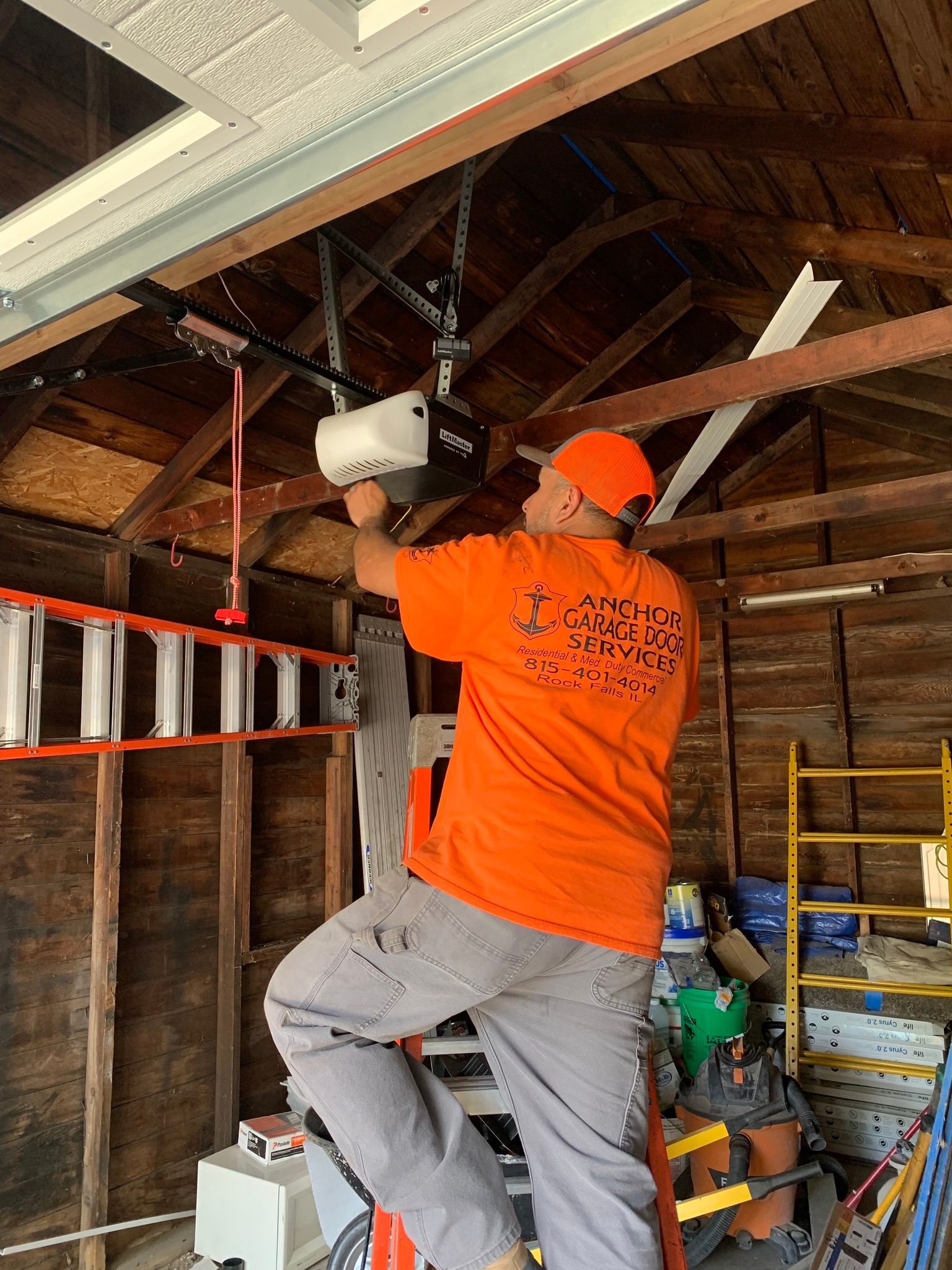 Man in orange shirt on ladder installing a garage door opener in a wooden garage.