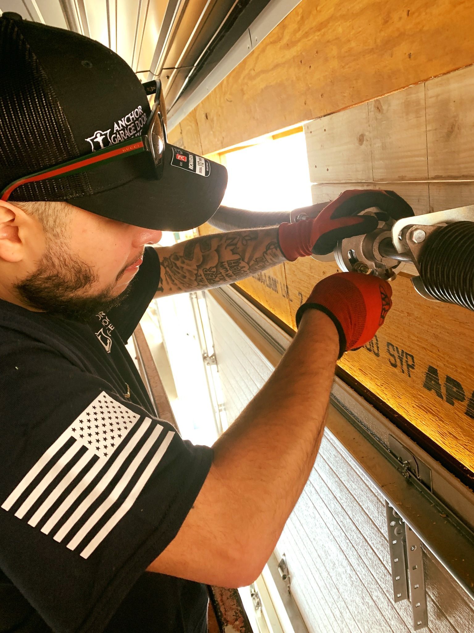 A man with tattoos, wearing a cap and gloves, repairs a garage door spring, indoors.