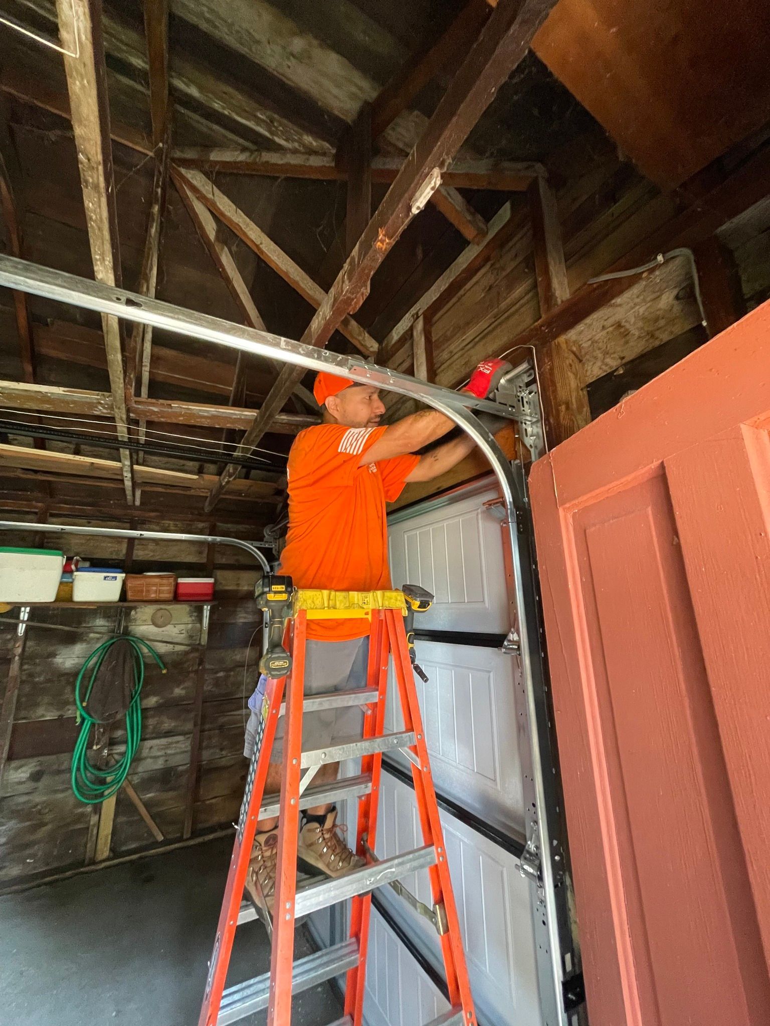 Man in orange shirt on ladder repairing garage door in old, wooden garage.