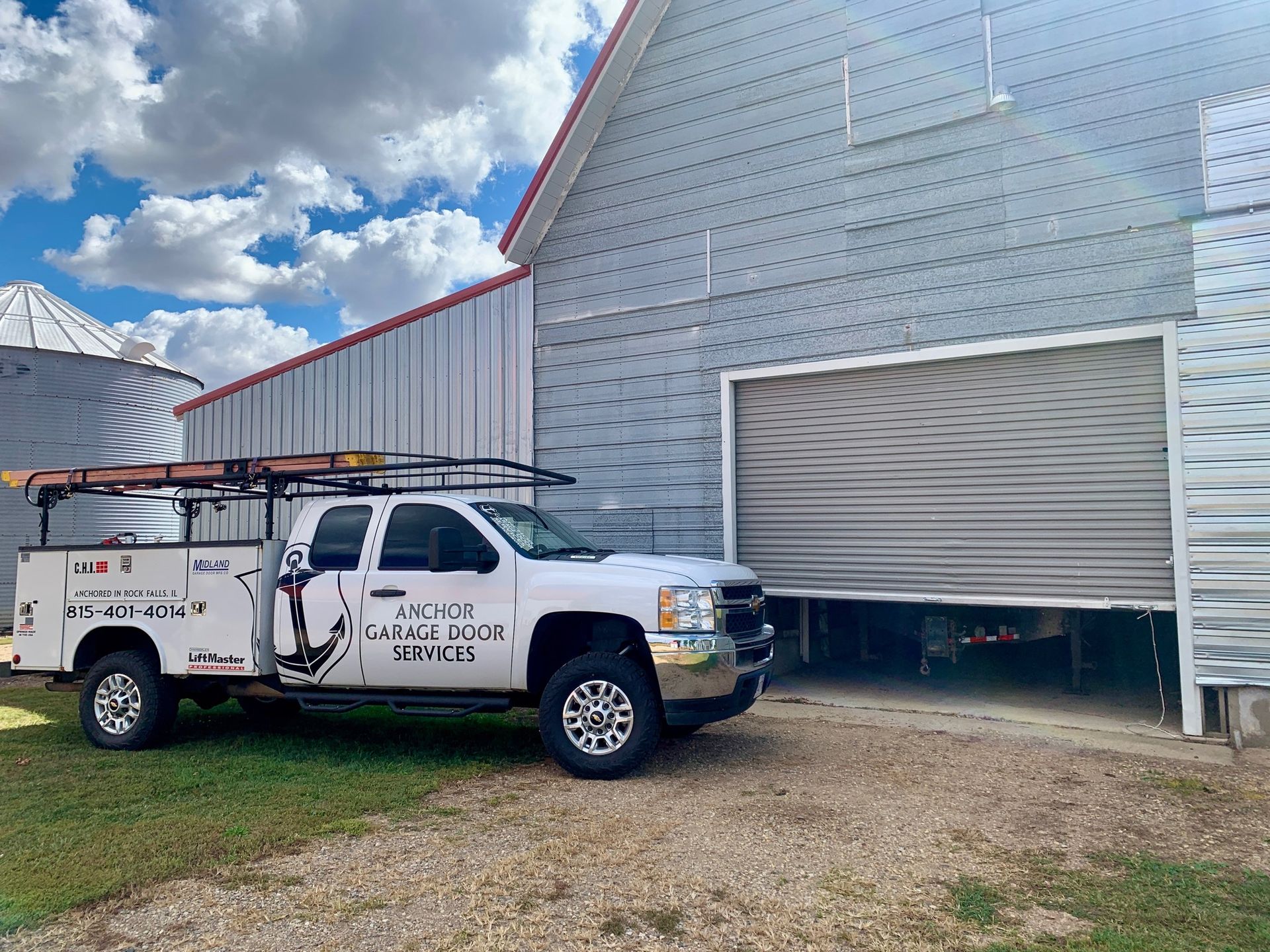 White truck with service logo parked in front of a grey garage door. Cloudy sky and building with a silo visible.