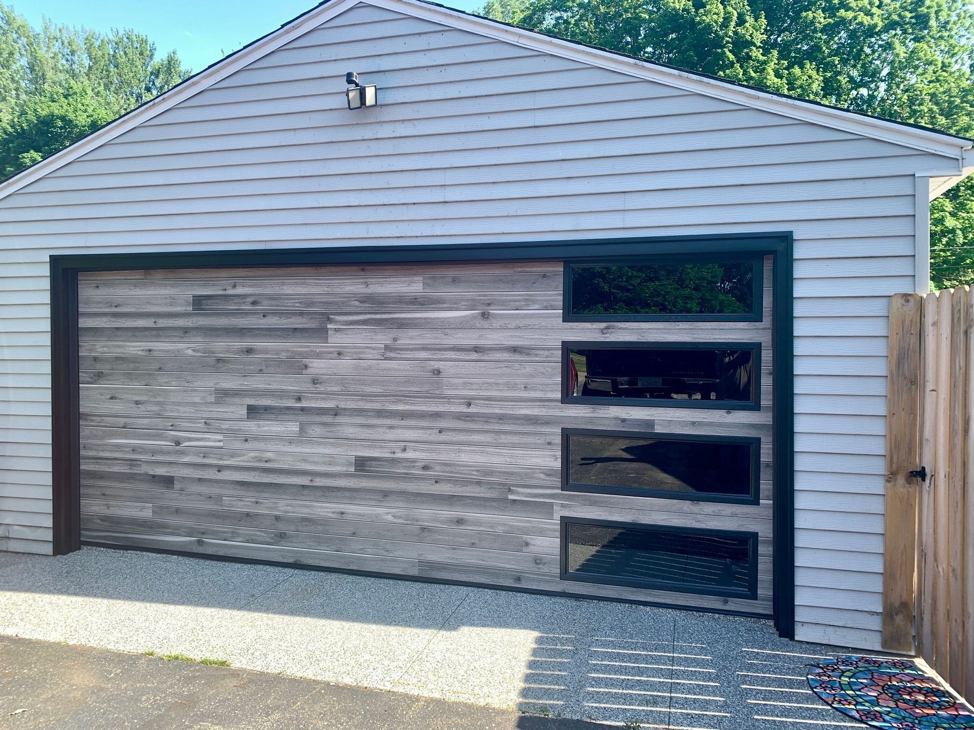 Garage with gray wood plank door, black frame, and glass windows; gravel driveway.