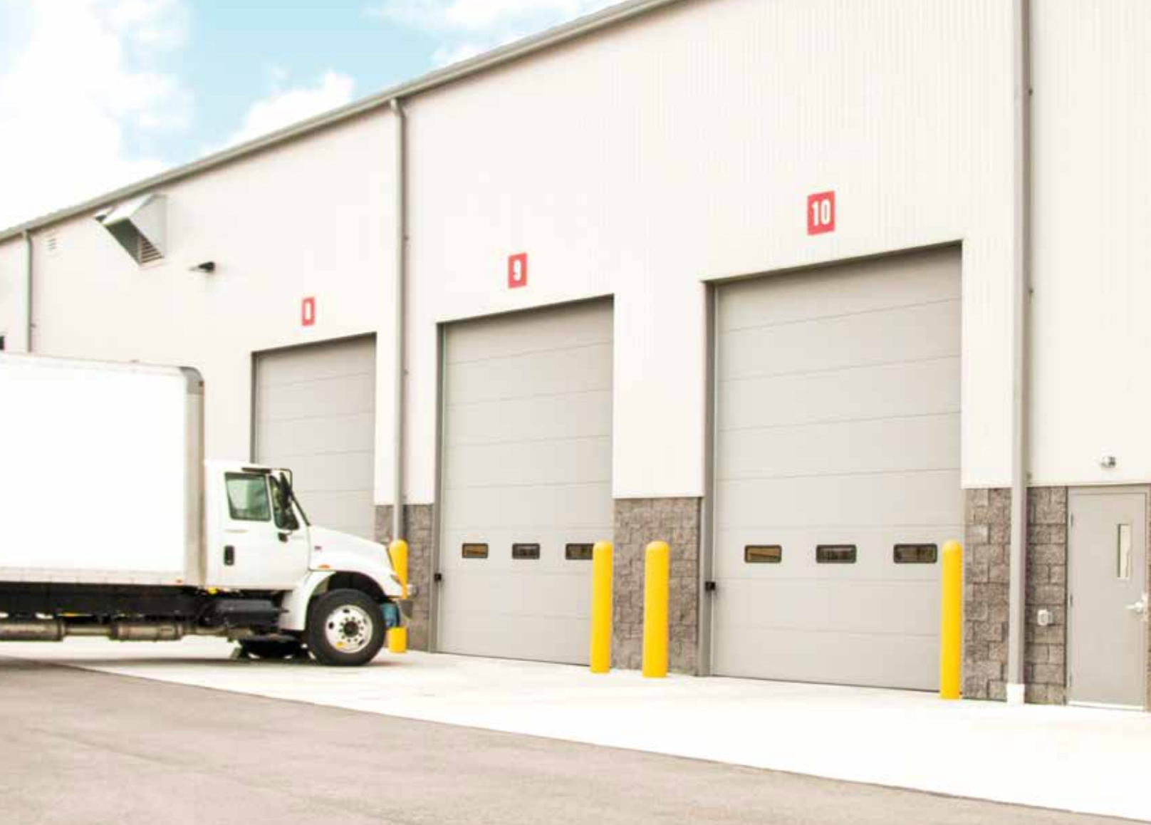 A truck backs into a loading dock at a white industrial building with three numbered doors.
