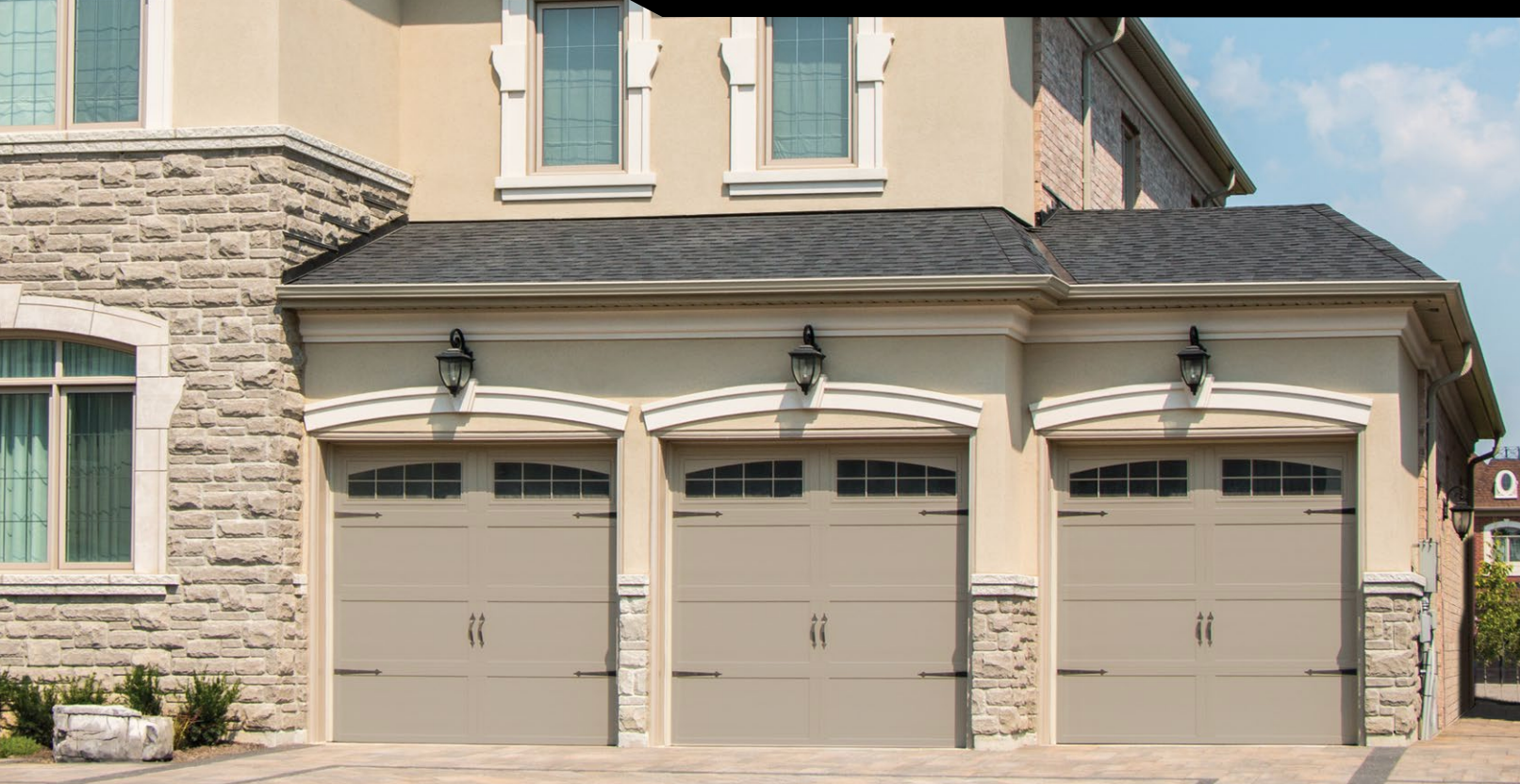 Three tan garage doors under a light beige stucco facade, with stone accents and black light fixtures.