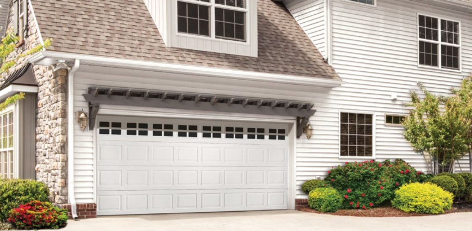 White garage door on a house with stone and white siding, surrounded by bushes and greenery.